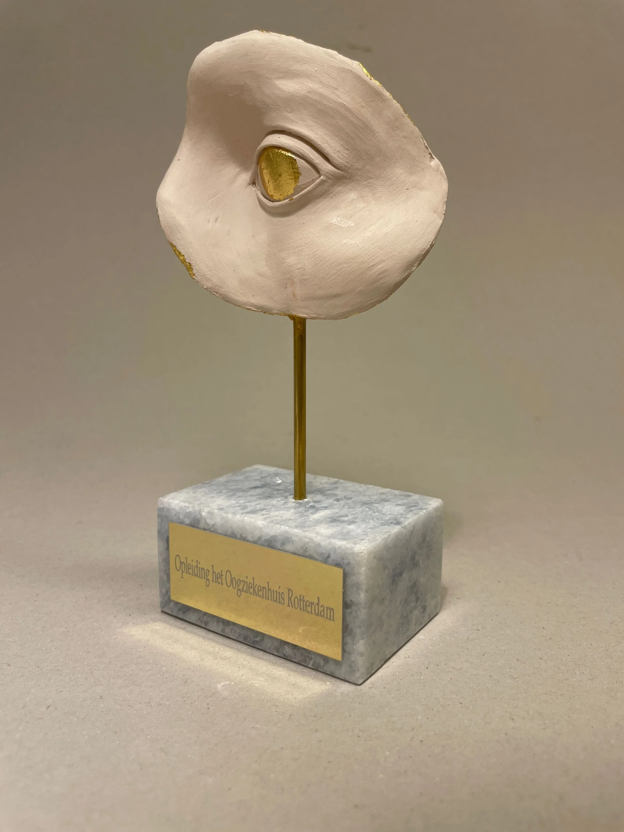 A sculpture of a human face in profile with a closed eye, painted white with gold accents around the eye and cheek, mounted on a marble base with a plaque that reads 'Opleiding het Oogziekenhuis Rotterdam'.