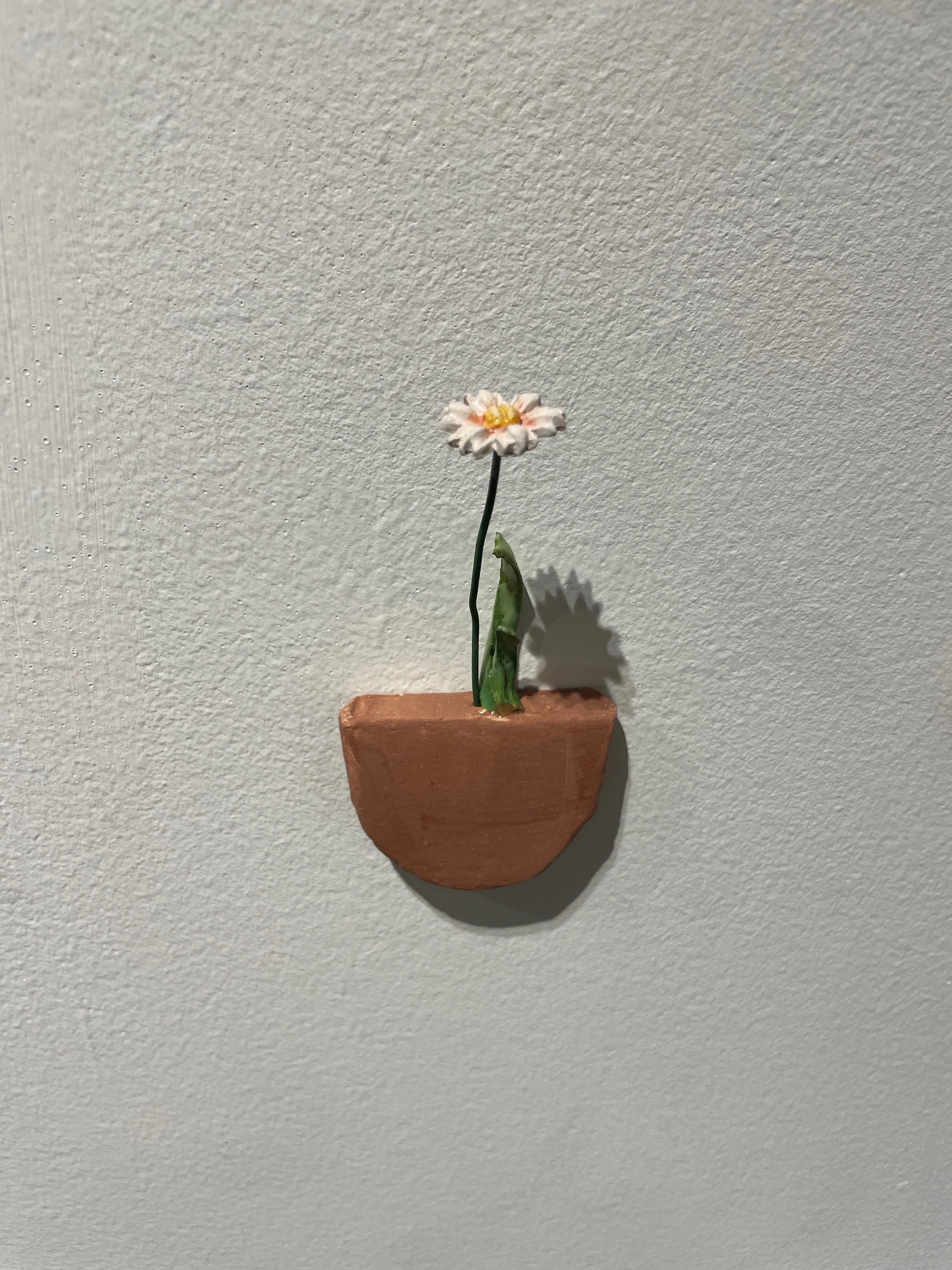 A small decorative flower sculpture with a pink and yellow flower in a terracotta pot mounted on a light-colored textured wall.