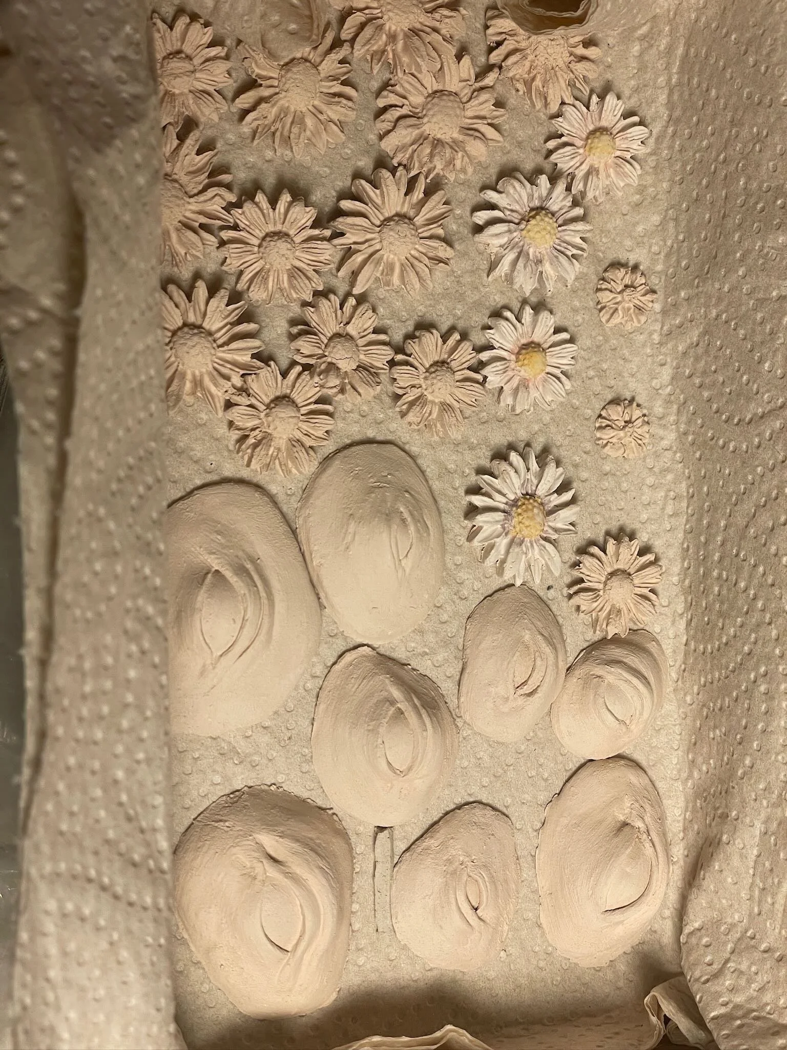 Clay sculptures of daisies and swirls on a textured surface, with the daisies on the upper part and the swirls on the lower part.