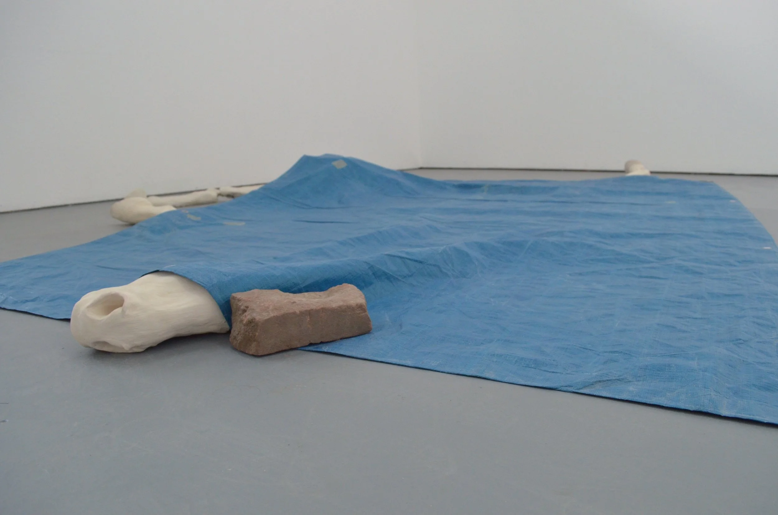 An art installation featuring a blue plastic sheet spread on the floor with white sculpture parts and a brick placed on it, against white walls.