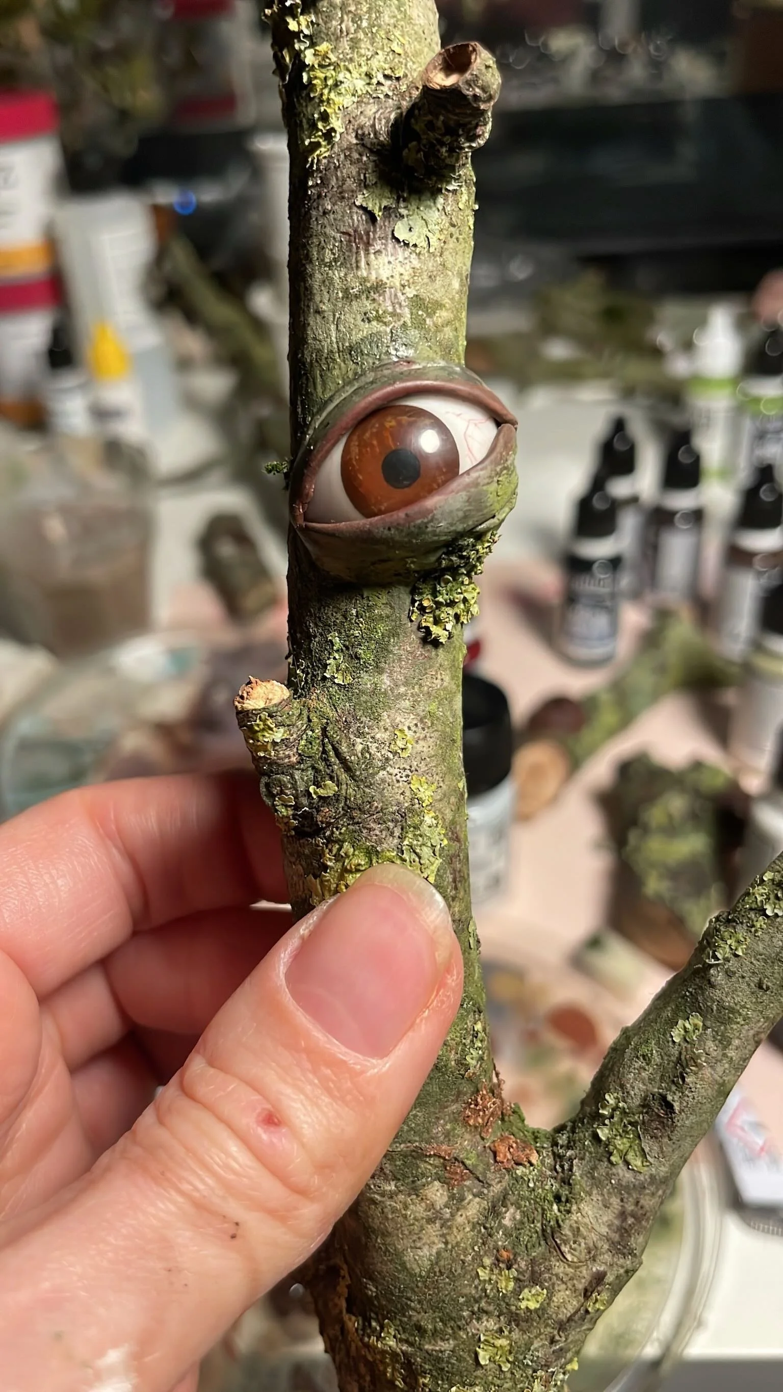 A small tree branch with a realistic eye sculpture embedded in the bark. The background shows art supplies and paint bottles.