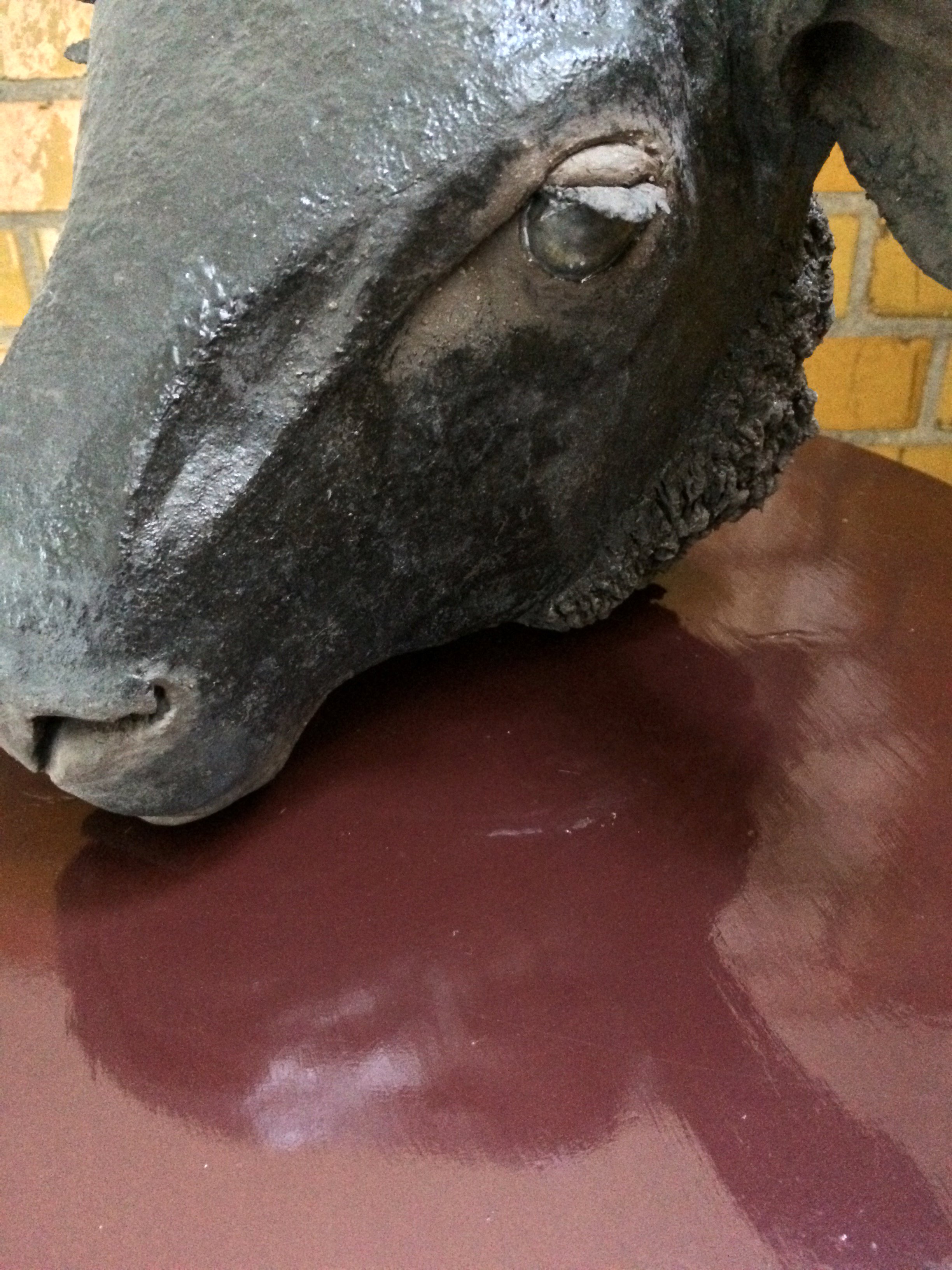 Close-up of a bronze or metal sculpture of a horse's head resting on a reddish-brown wooden surface, with a brick wall in the background.