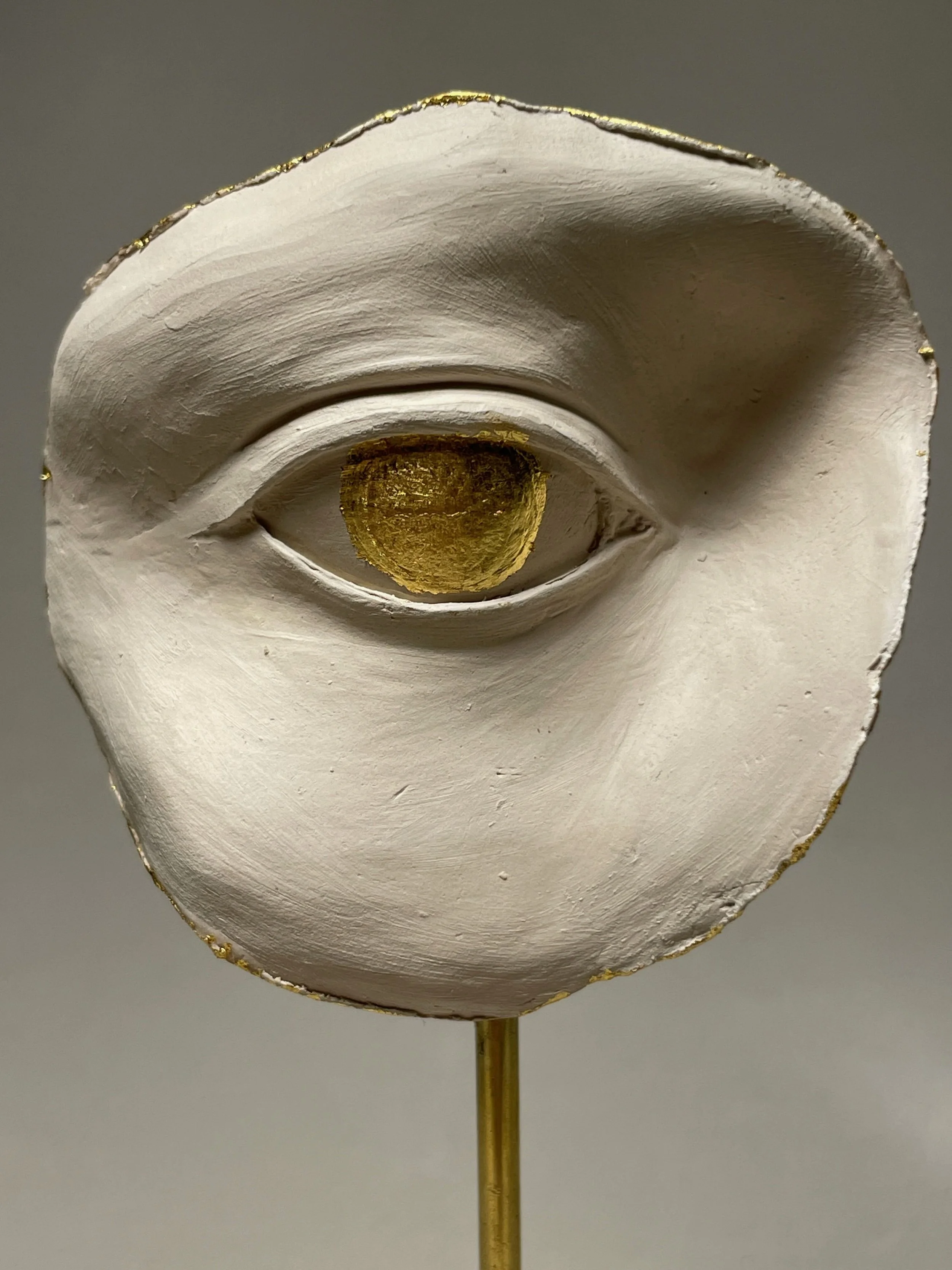 Close-up of a white, textured sculpture of a face with an eye in the center, surrounded by a shell-like structure with gold accents, mounted on a gold stand.