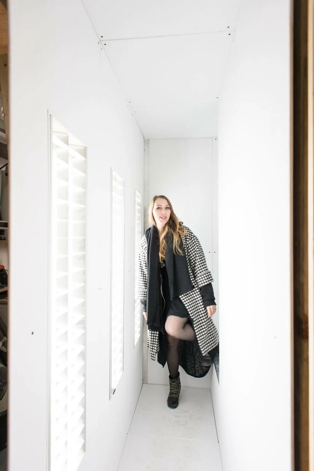 A woman standing in a small, white, enclosed space with vents on the wall, wearing a patterned coat, black scarf, black dress, tights, and boots, posing with one leg raised.