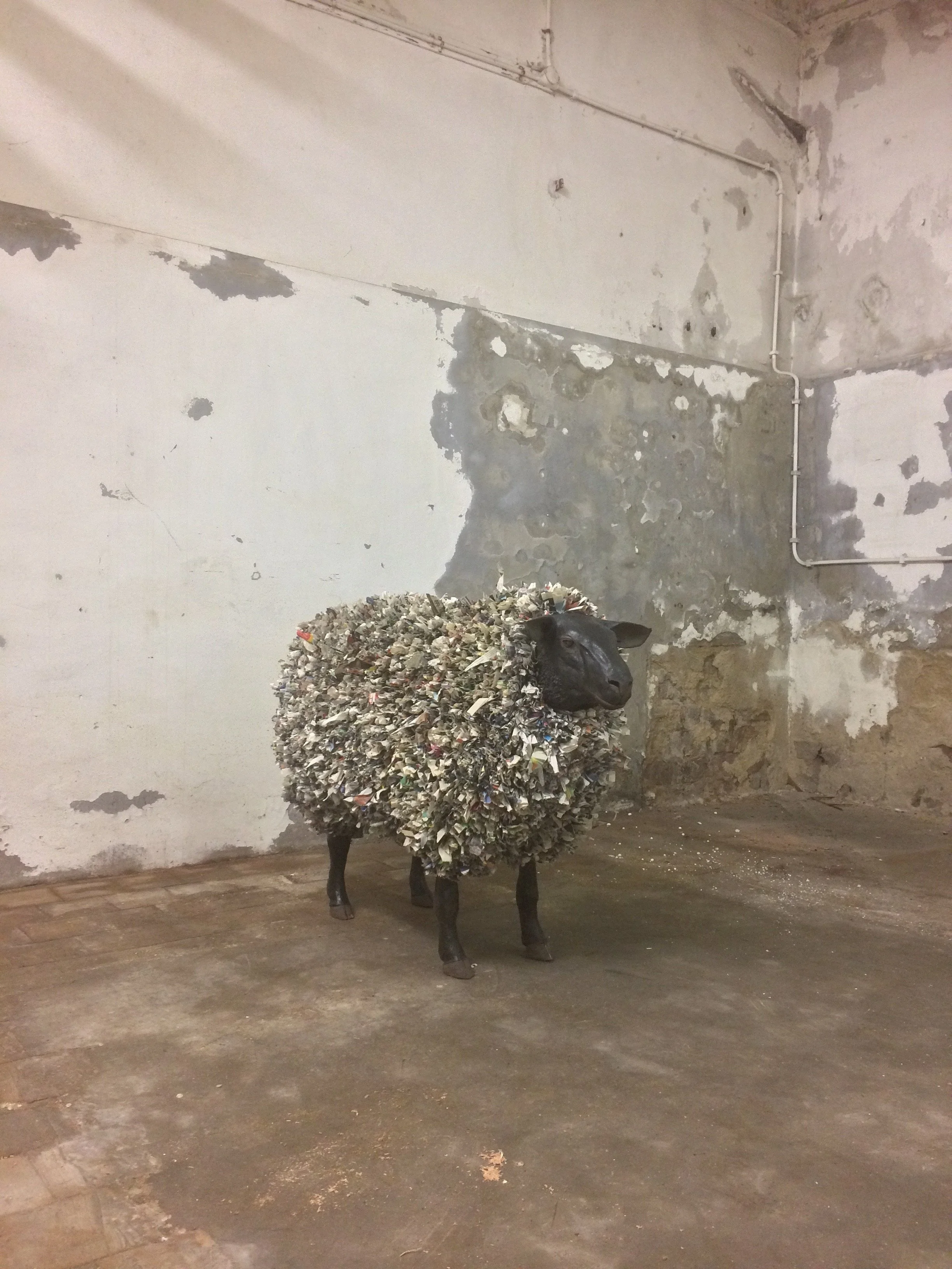 A sculpture of a sheep with a body made of shredded paper or plastic, standing on a concrete floor in front of a weathered, peeling wall with exposed pipes.