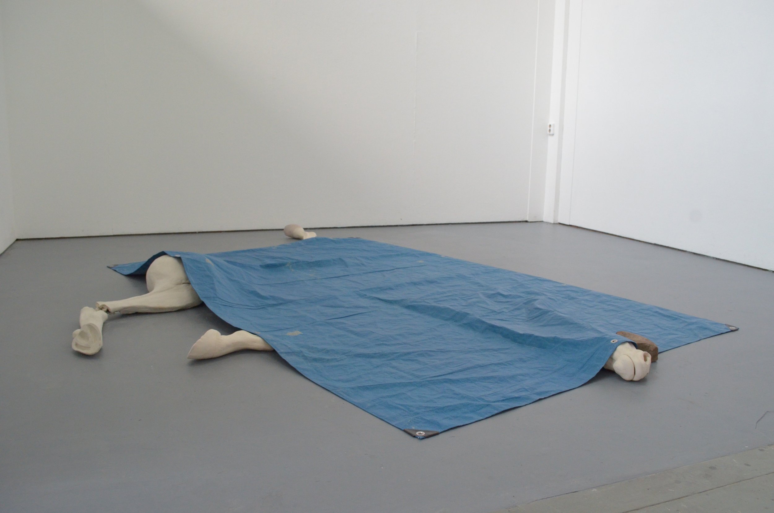 An art installation featuring a human figure lying face down on the floor, covered with a blue tarp, in a minimal white gallery space.