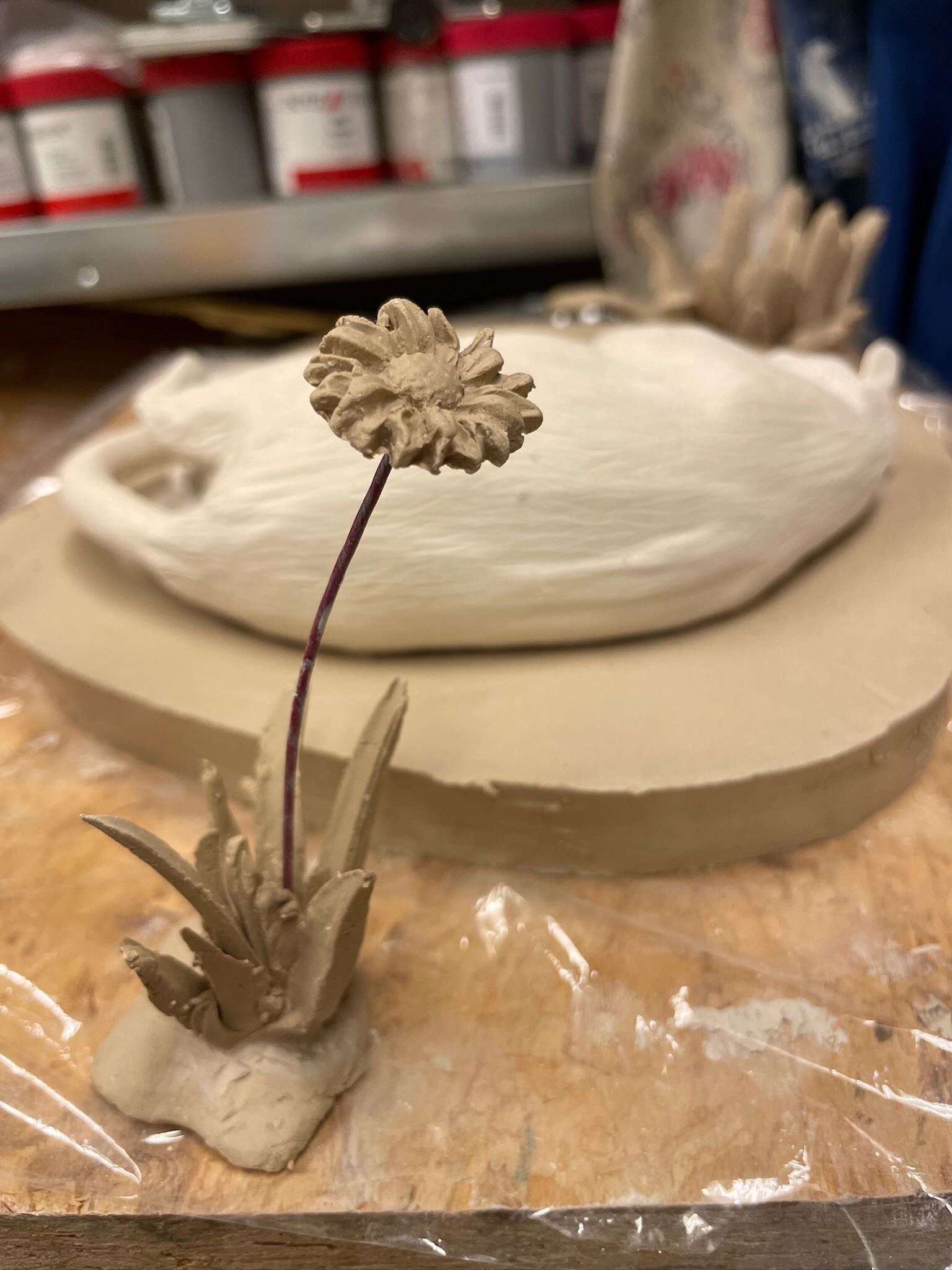Unfinished ceramic sculpture of a small plant with a flower, attached to a base, placed on a wooden workspace with a plaster mold and art supplies in the background.