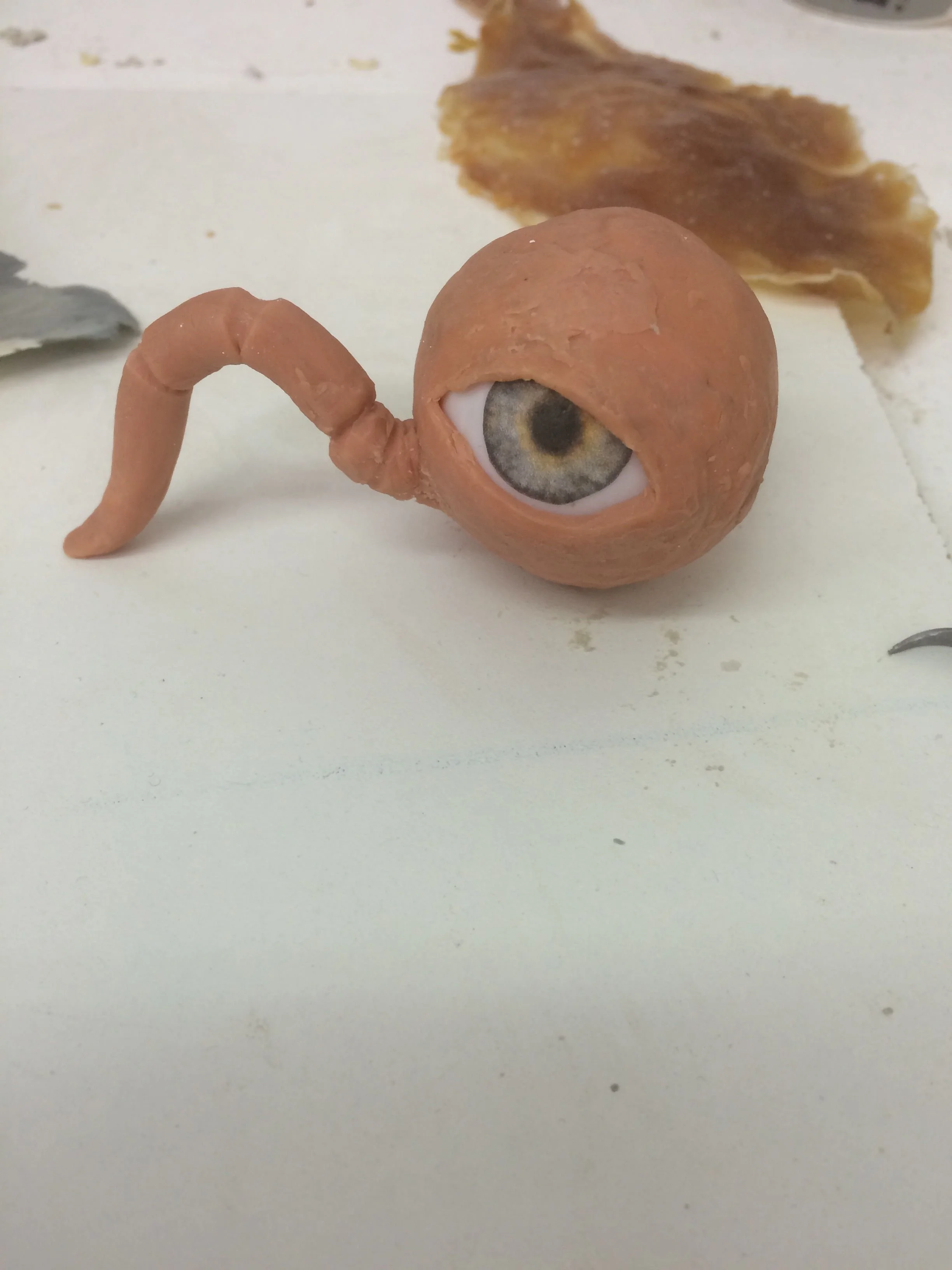 A rubber object with a human eye printed on it, attached to a flexible tube, resembling a creature with an eye for a head and a tail, on a white surface with some pieces of bread and a vegetable in the background.