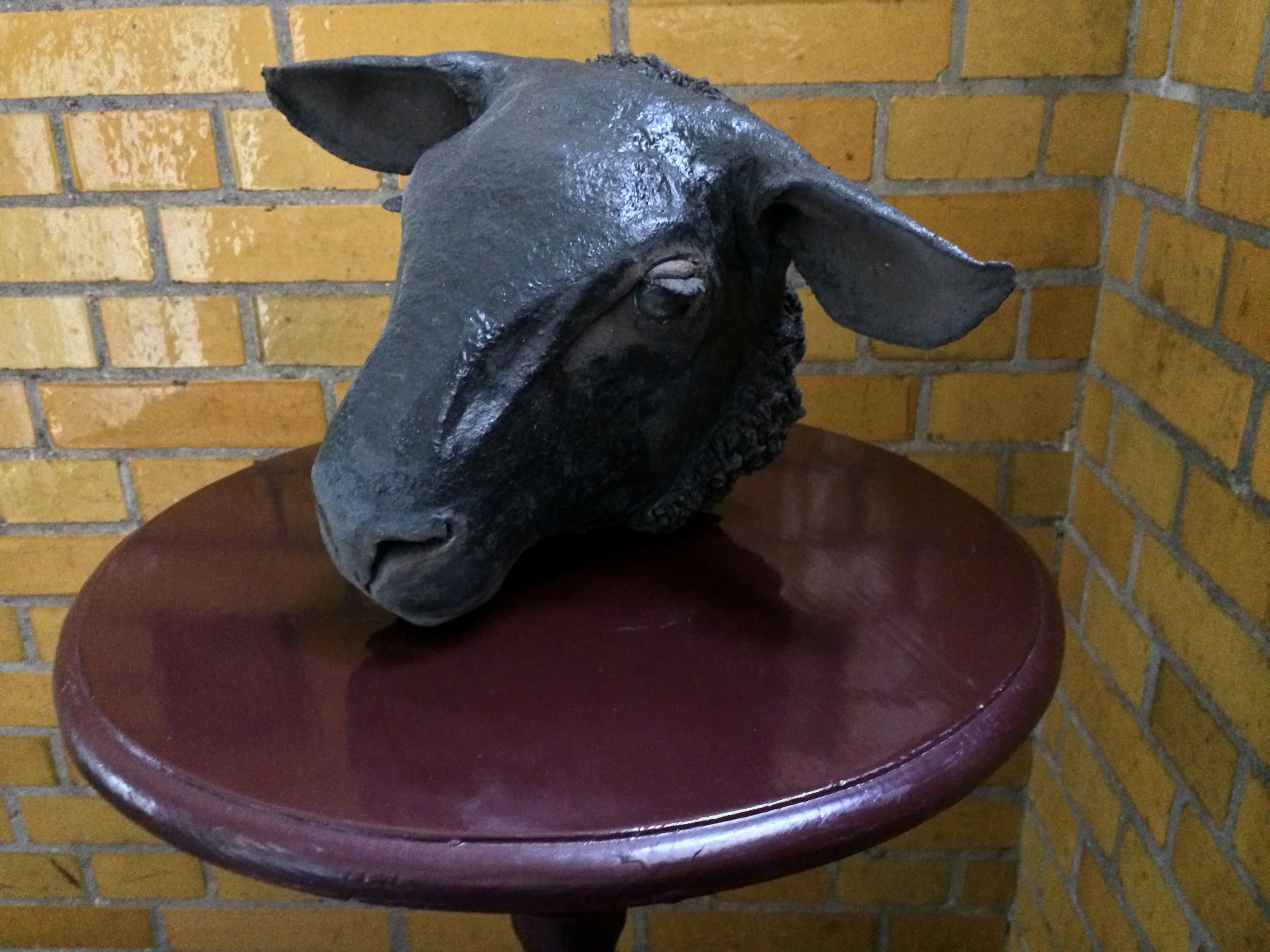 A horse head sculpture on a dark wooden table, against a yellow brick wall background.