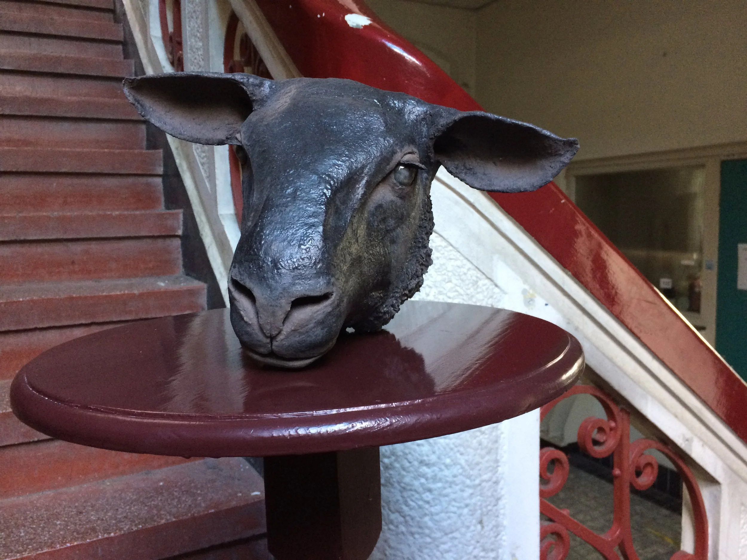 Bronze sculpture of a goat's head on a wooden stand, positioned at a staircase in an indoor setting.