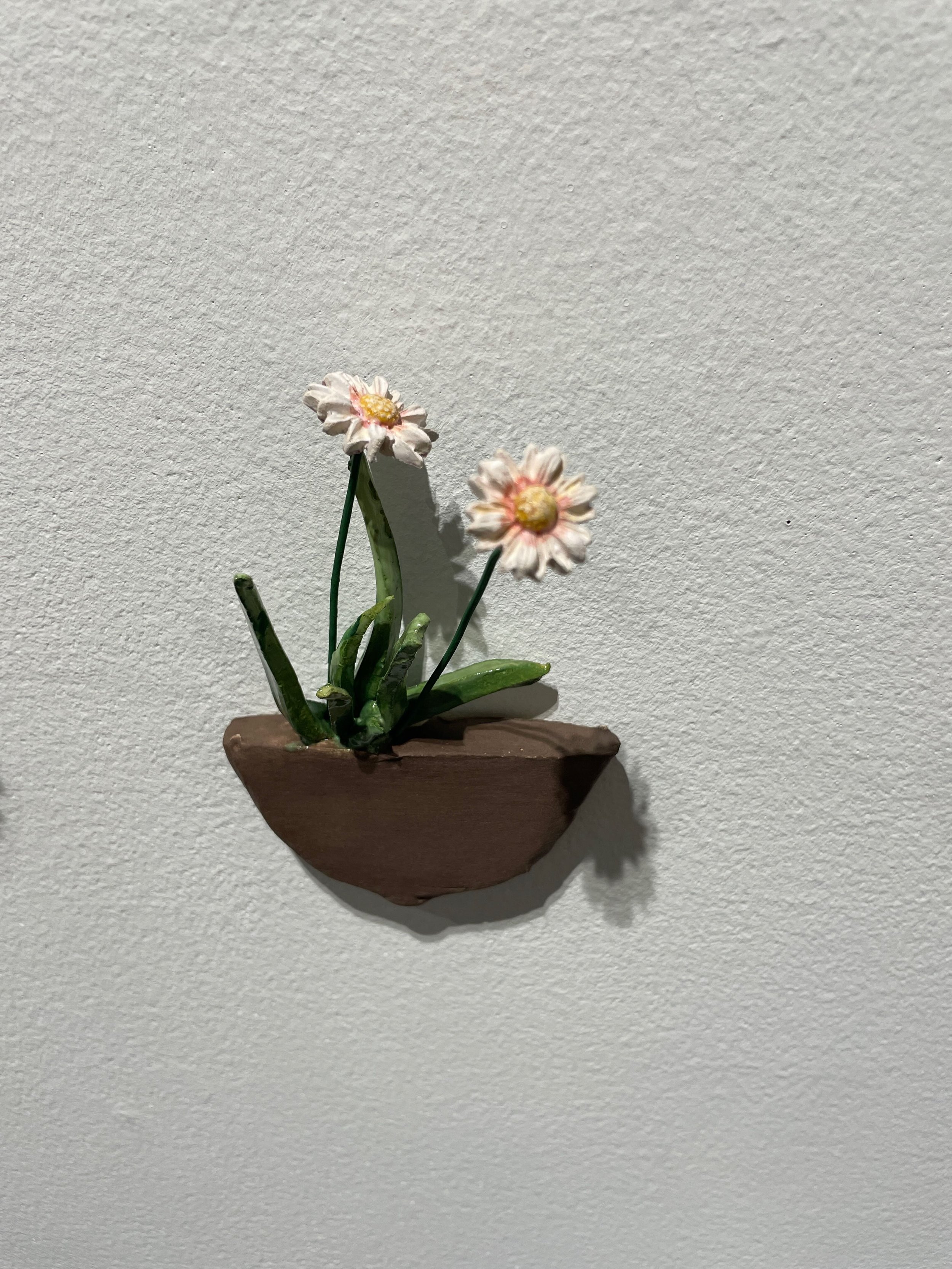 Small handcrafted clay flower pot with two white and pink painted flowers and green leaves, mounted on a white wall.