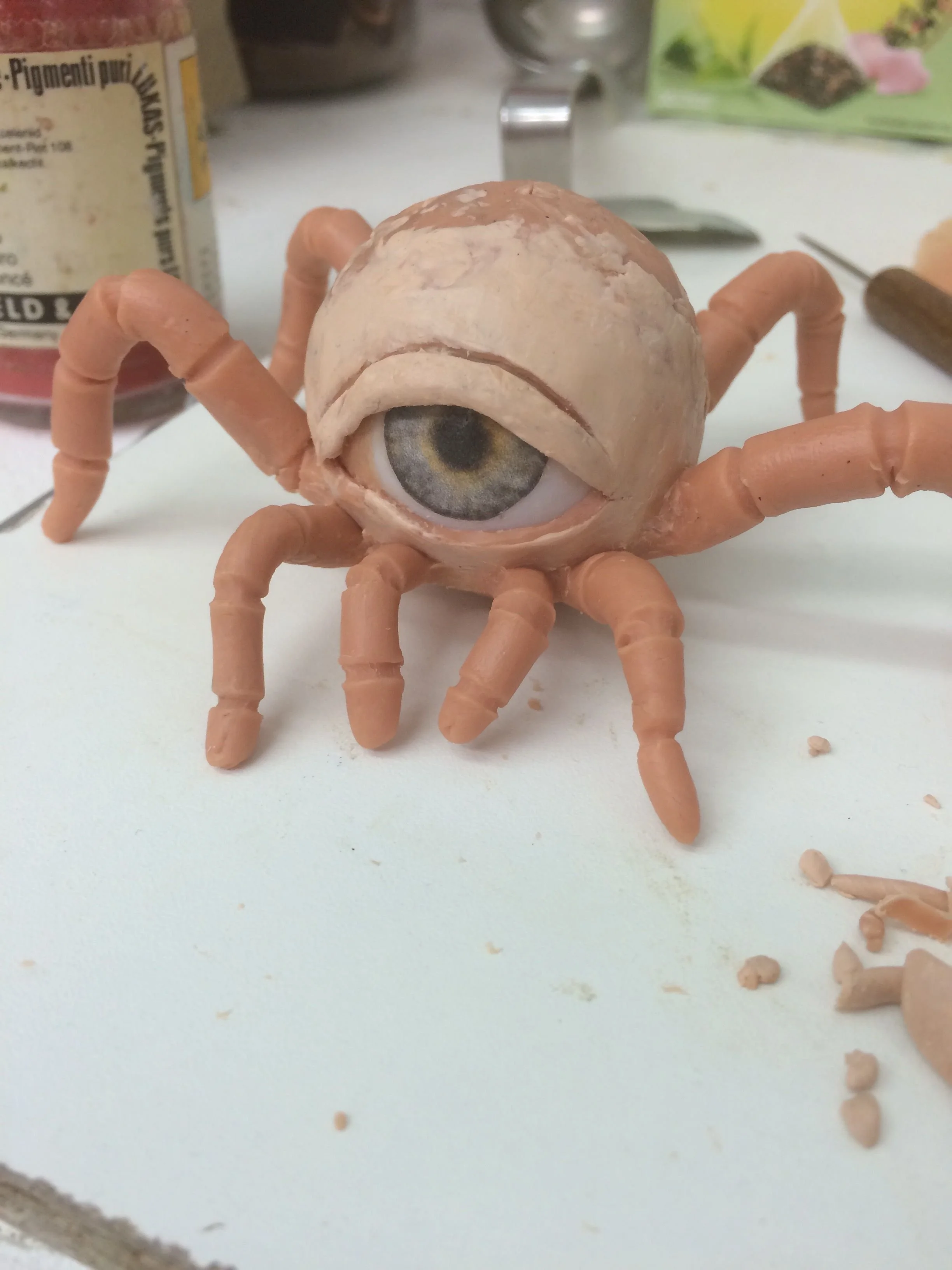 A craft project resembling a spider with a large central eye featuring a human eye in the middle, surrounded by clay legs on a white surface with various crafting tools and materials in the background.