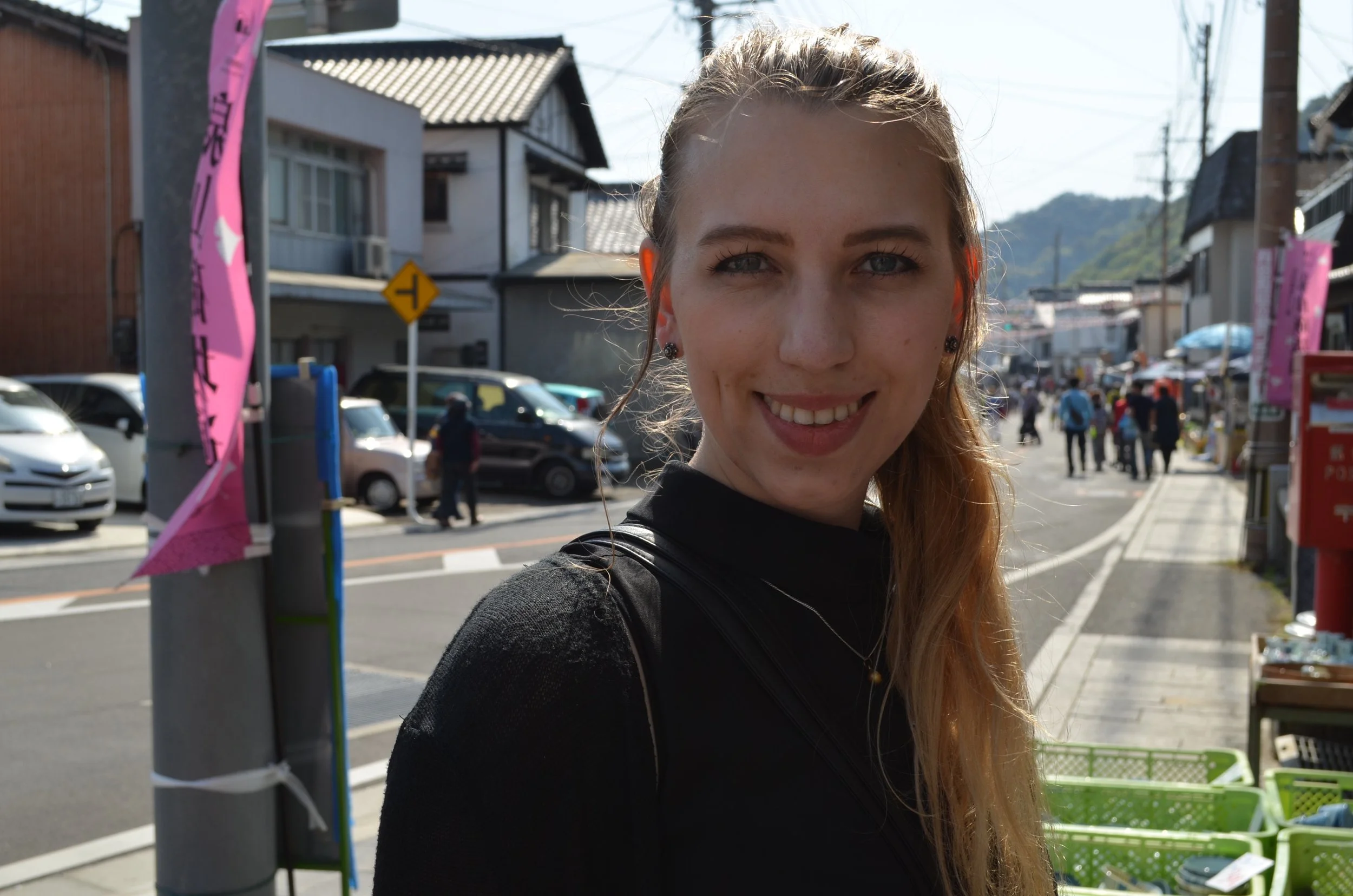 Young woman smiling outdoors on a sunny day with a street and shops in the background.