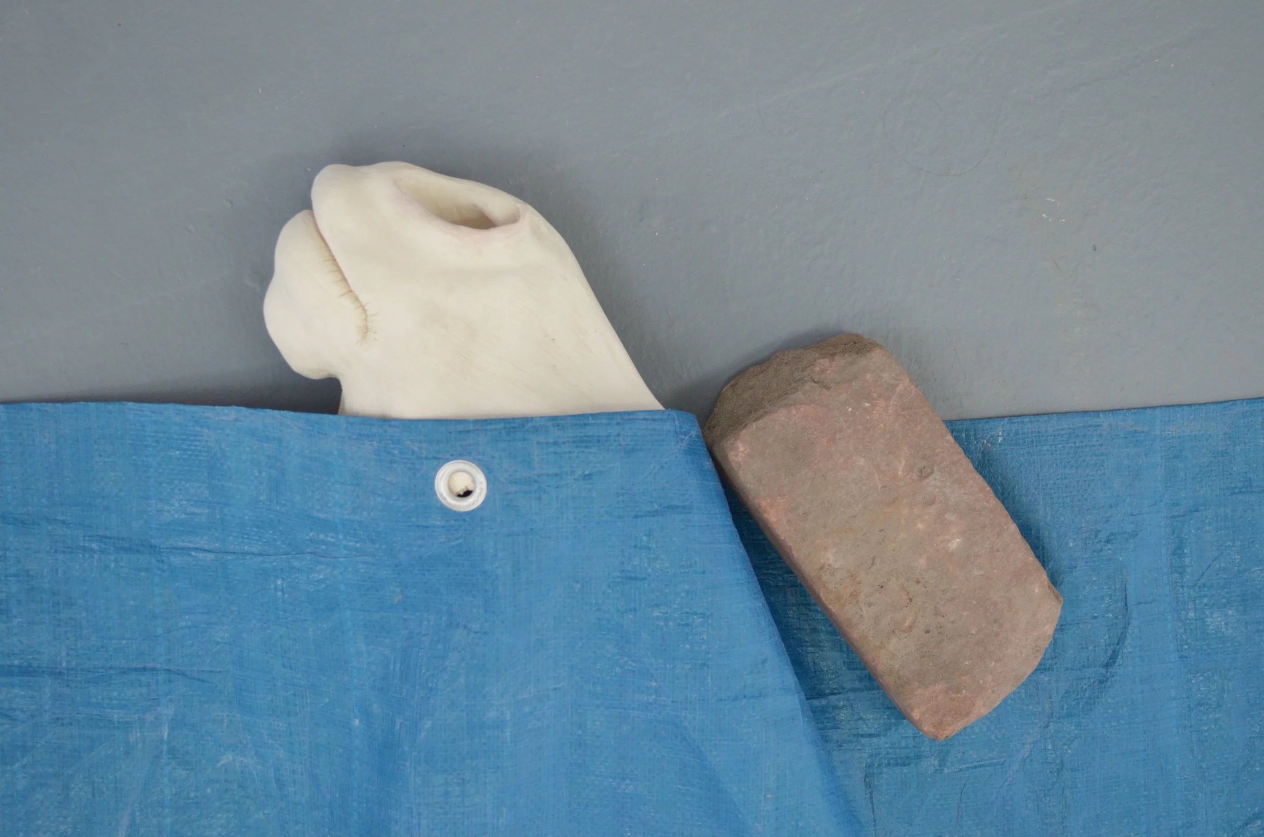 A white animal skull, possibly a horse skull, partially covered by a blue fabric with a white button, resting against a gray wall beside a reddish-brown brick.