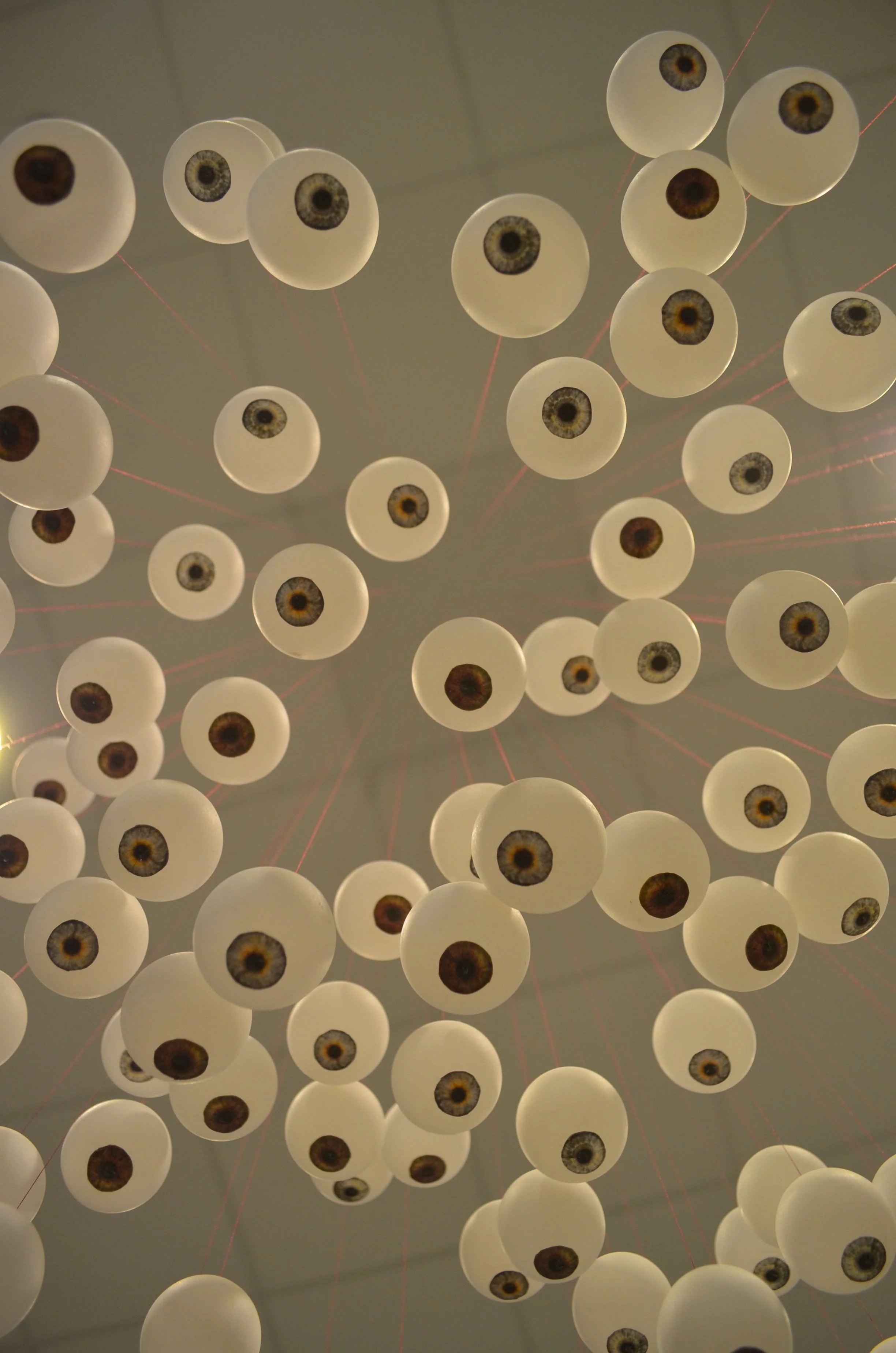 A modern chandelier with numerous white spherical lights, each featuring an eye-like design with an iris and pupil, hanging from thin red wires.