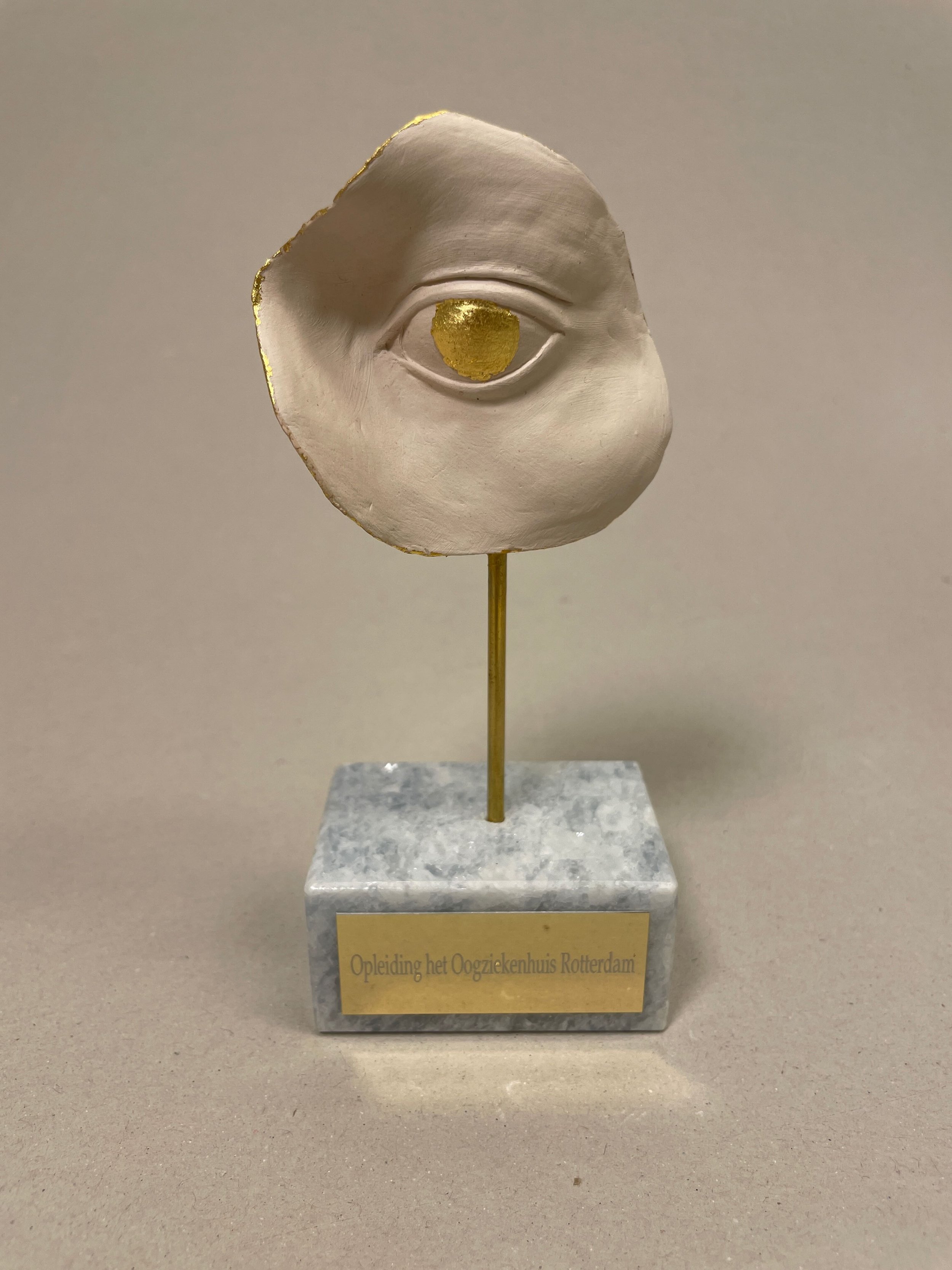 A sculpture of a human eye on a stand with a marble base. The sculpture is primarily white with gold accents on the eye and edges. There is a gold plaque on the base with Dutch text.