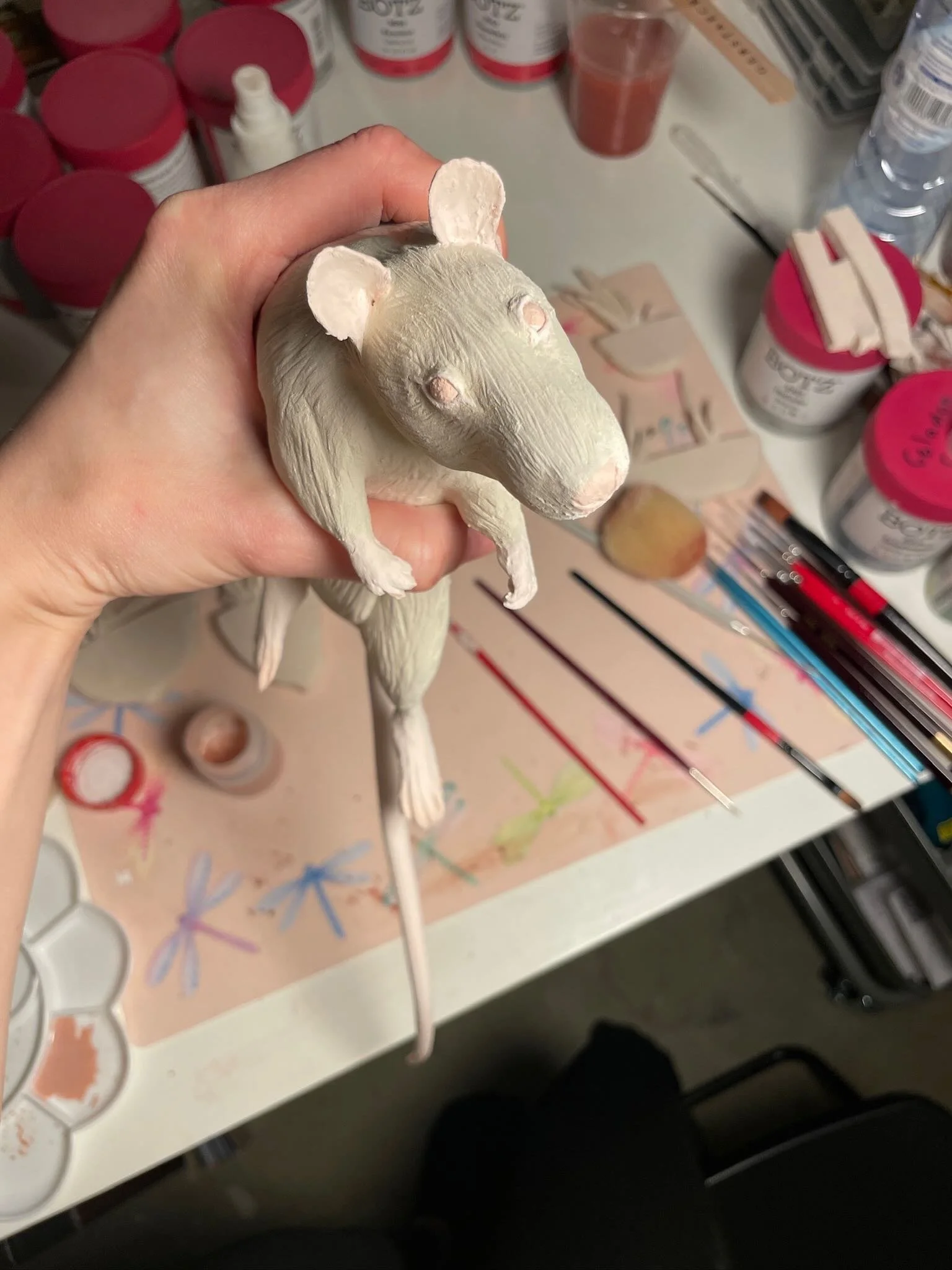 Hand holding a sculpted and painted animal figure with the body of a mouse or rat and the head of a dog, surrounded by art supplies on a workspace.