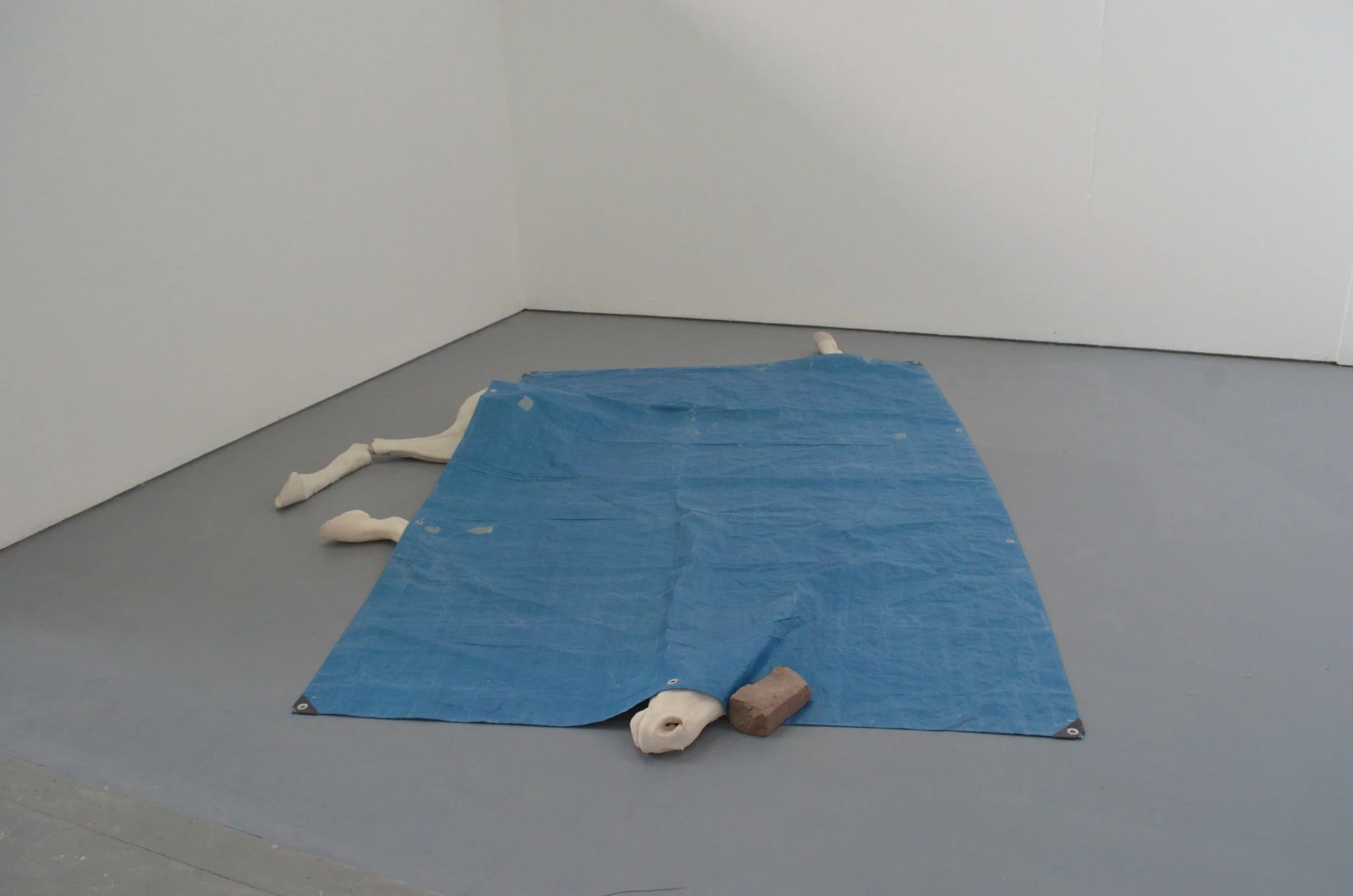 An art installation depicting a white, mannequin-like figure lying flat on a gray floor, covered partially with a blue tarp, in a corner of a white room.
