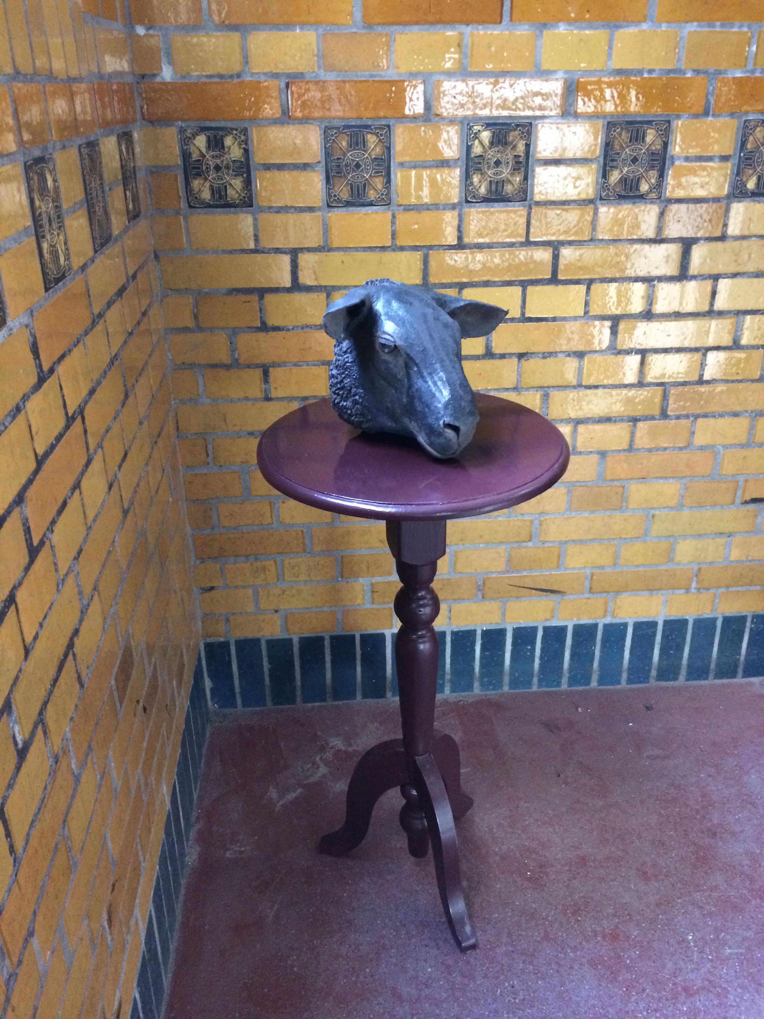 A black sculpture of a goat's head on a small round wooden table against a brick wall.