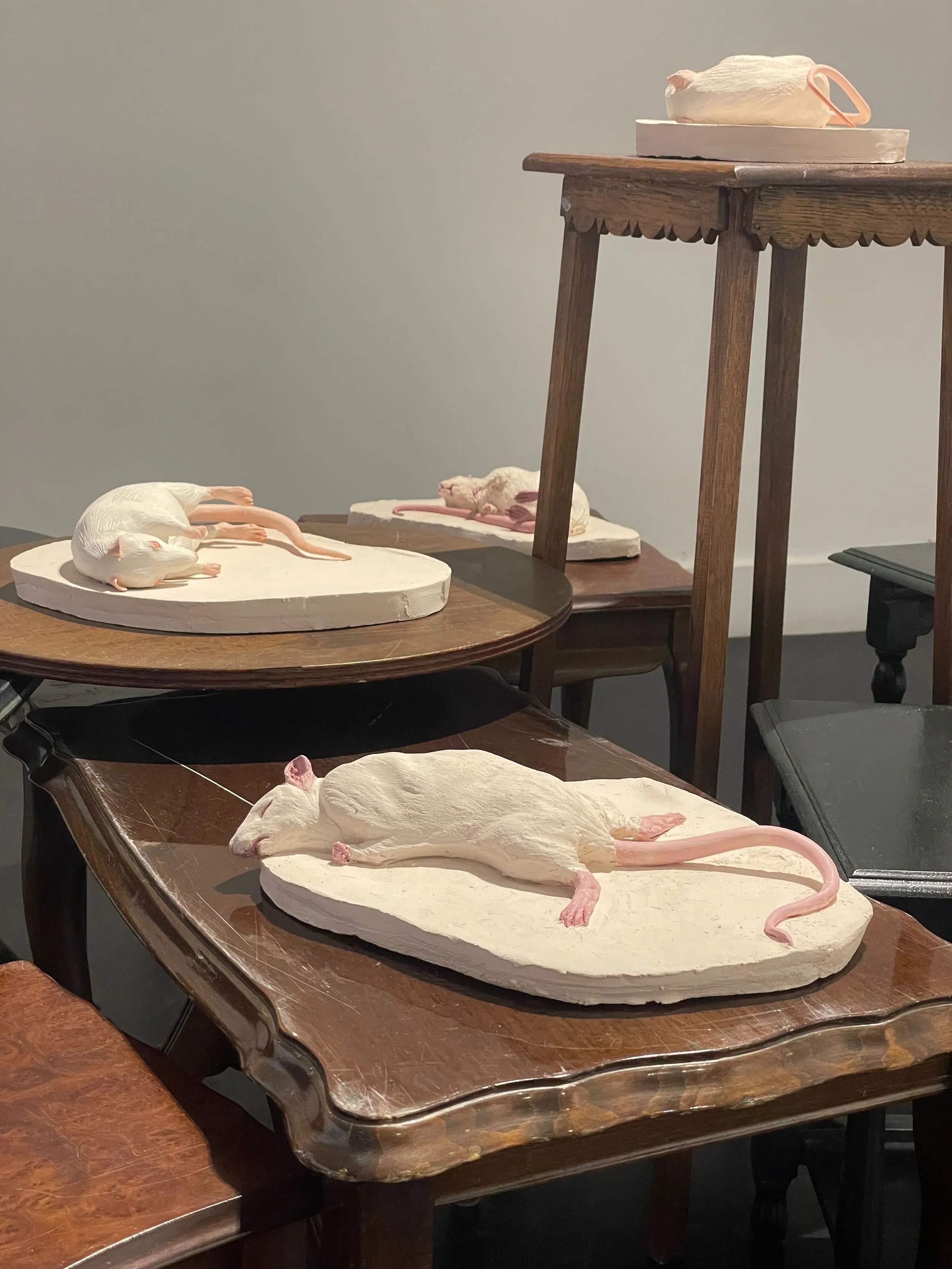 Artistic sculptures of mice lying on white bases, displayed on wooden tables.