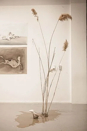 Dried tall grass arranged in a vase with a small bird sculpture on the floor in front of it.
