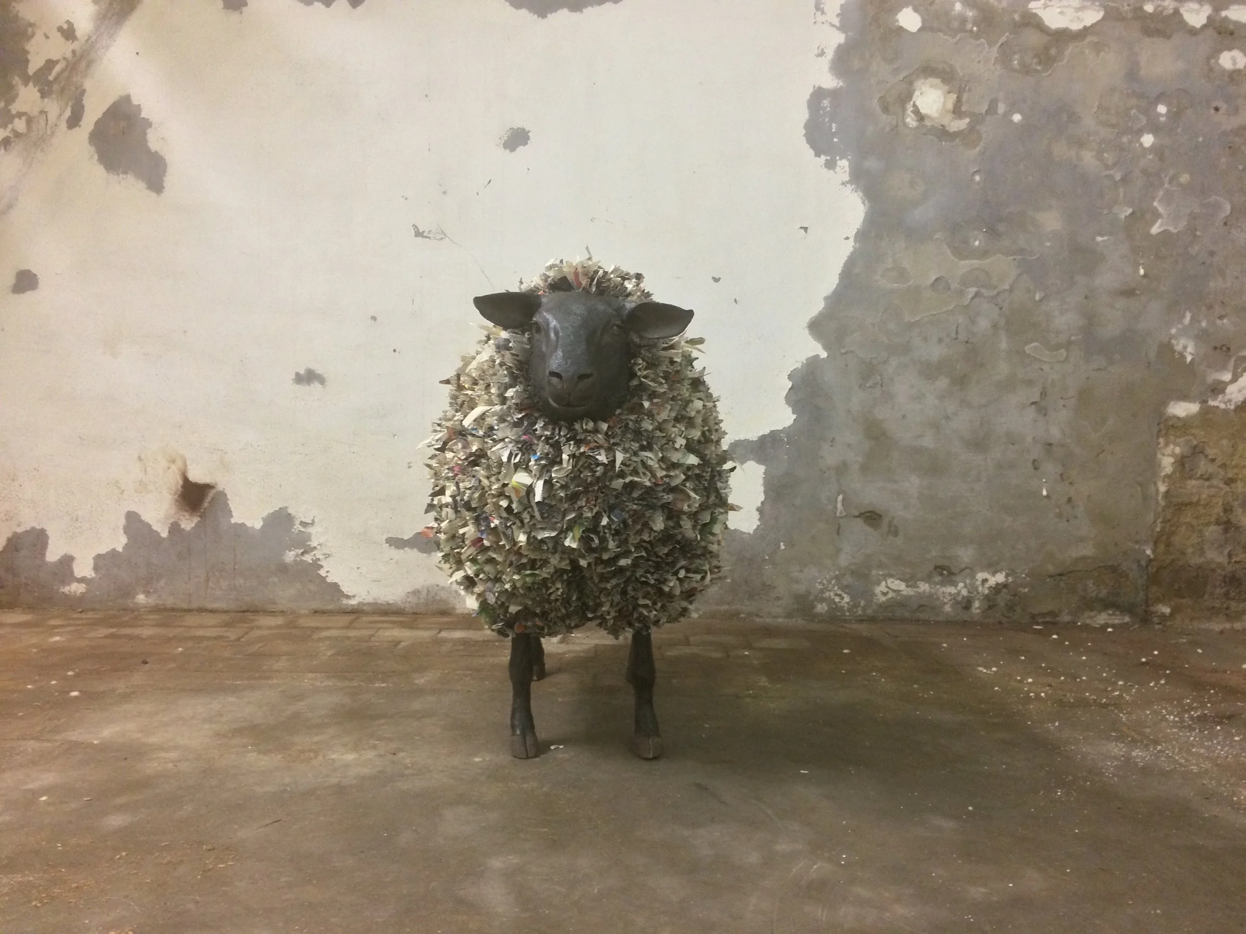 A sheep sculpture with a body made of shredded paper and a painted black head, standing on a concrete floor against a weathered wall.