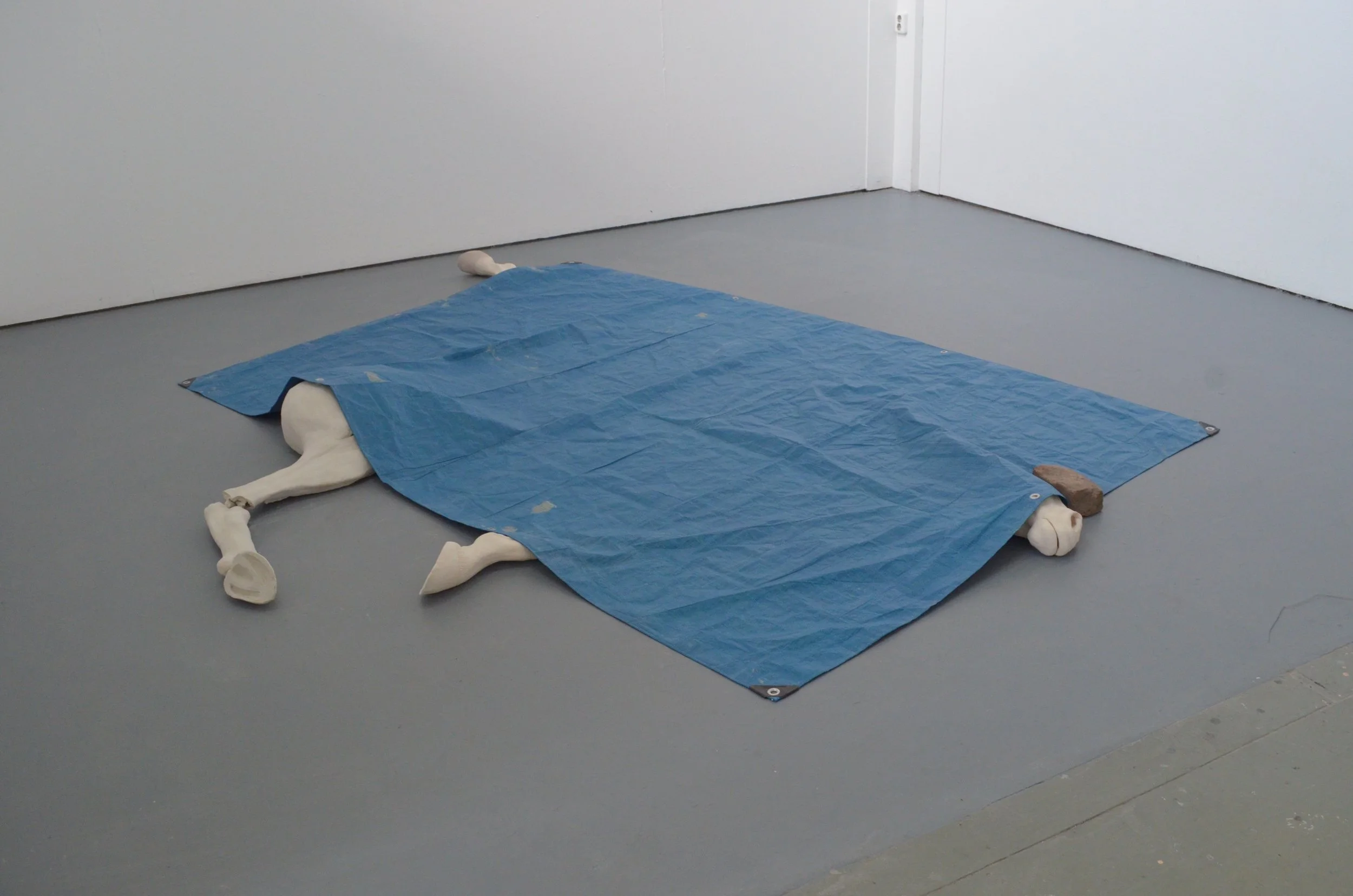 A mannequin lying face down on a blue sheet on a gray floor in an empty white-walled room.