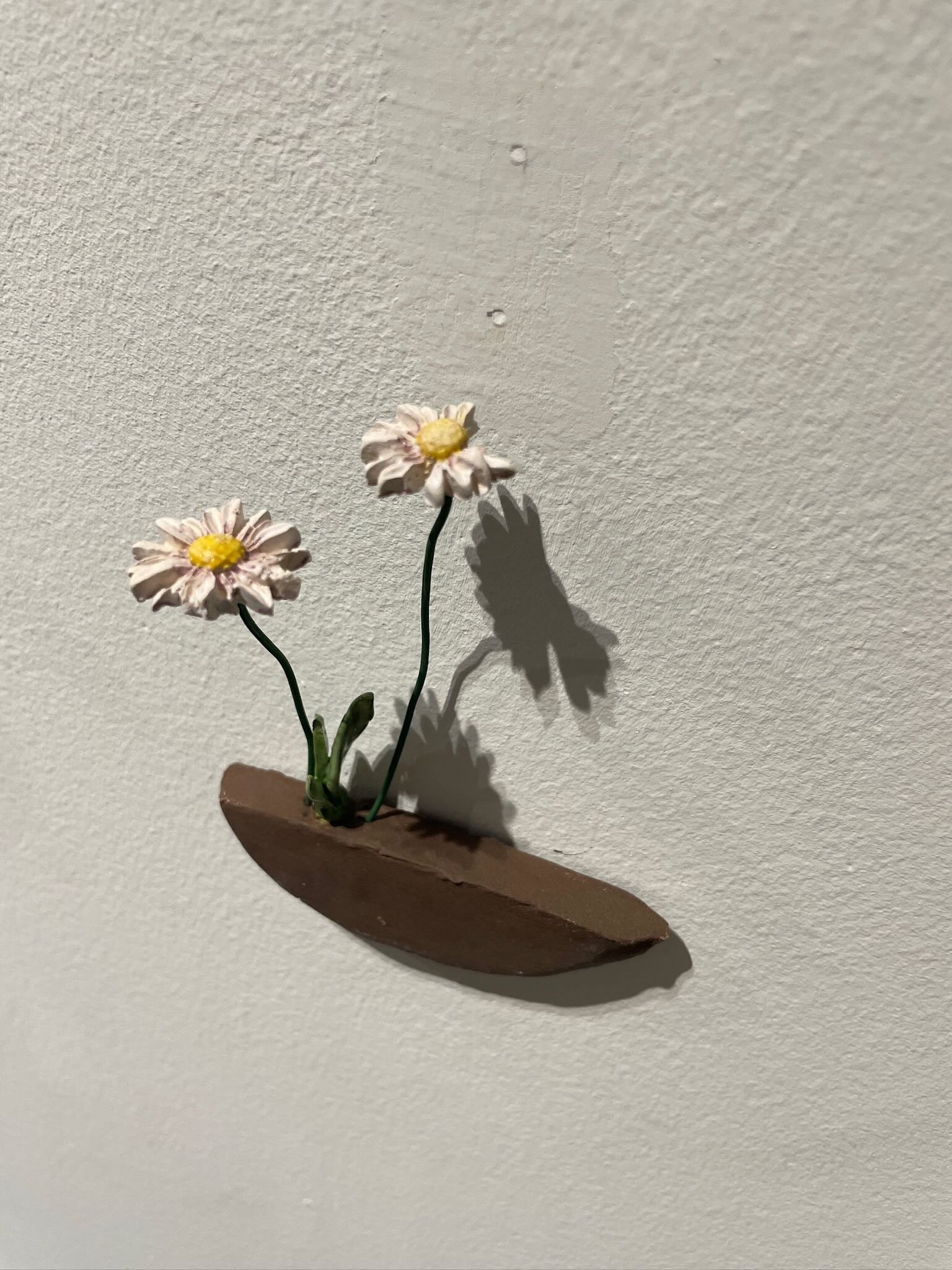 Artificial flowers resembling daisies with pink petals and yellow centers, mounted on a small piece of brown wood, casting shadows on a white wall.