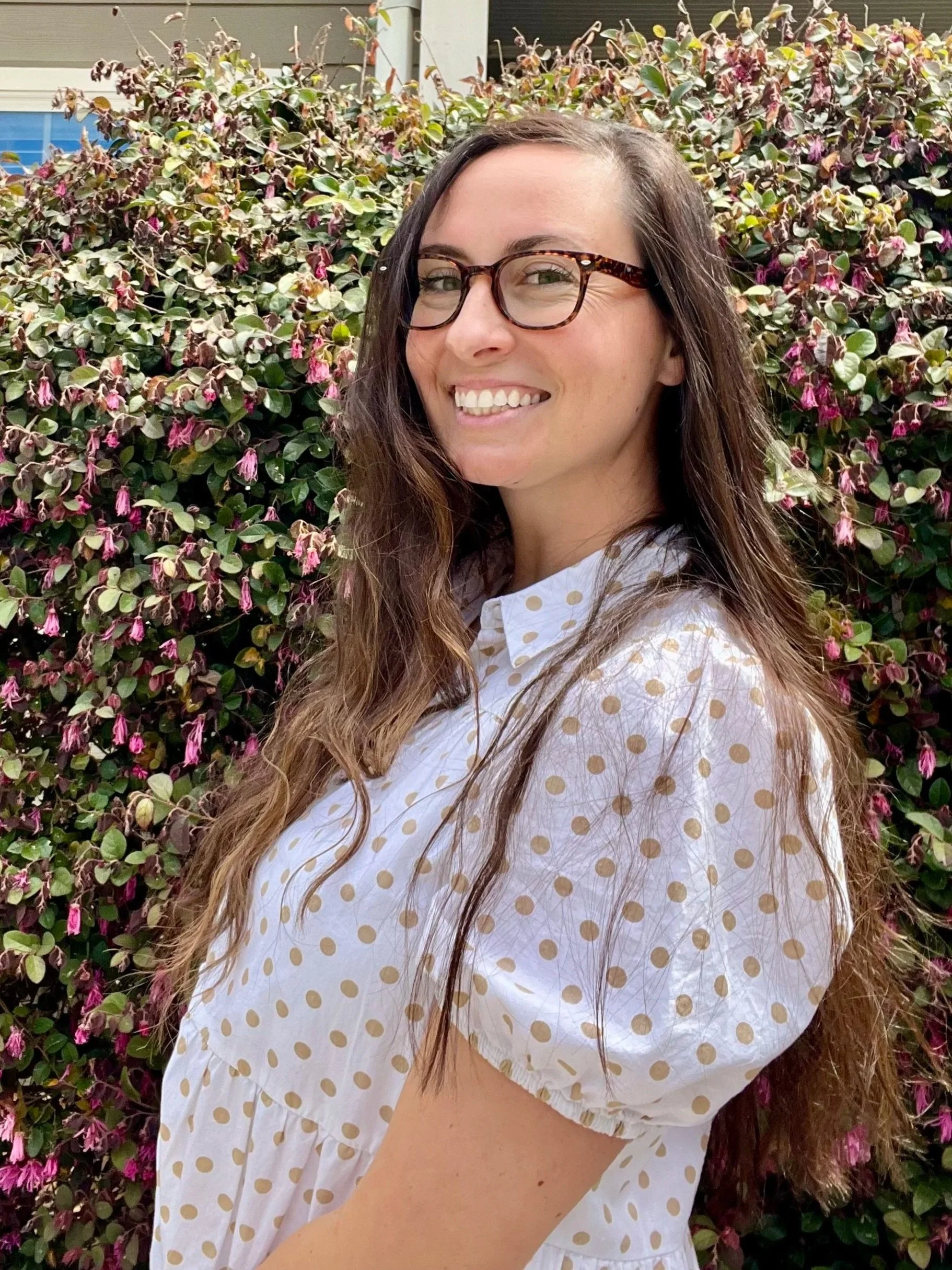 A woman with long, wavy brown hair, wearing tortoiseshell glasses and a white polka-dot blouse, smiling in front of a bush with pink and purple flowers.