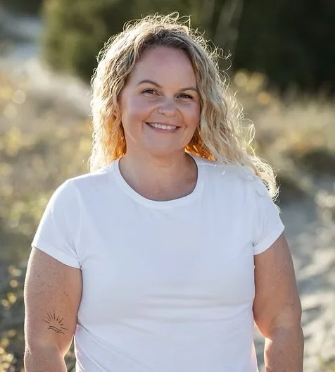 A woman with curly blonde hair smiling outdoors in a white t-shirt, with trees and sunlight in the background.