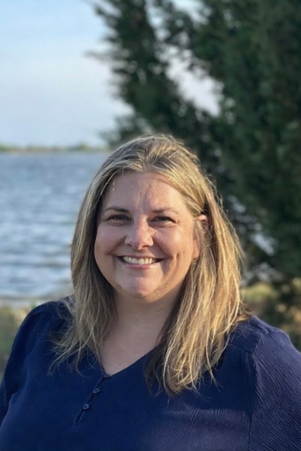 A woman with shoulder-length blonde hair smiling outdoors near a body of water and tall trees.