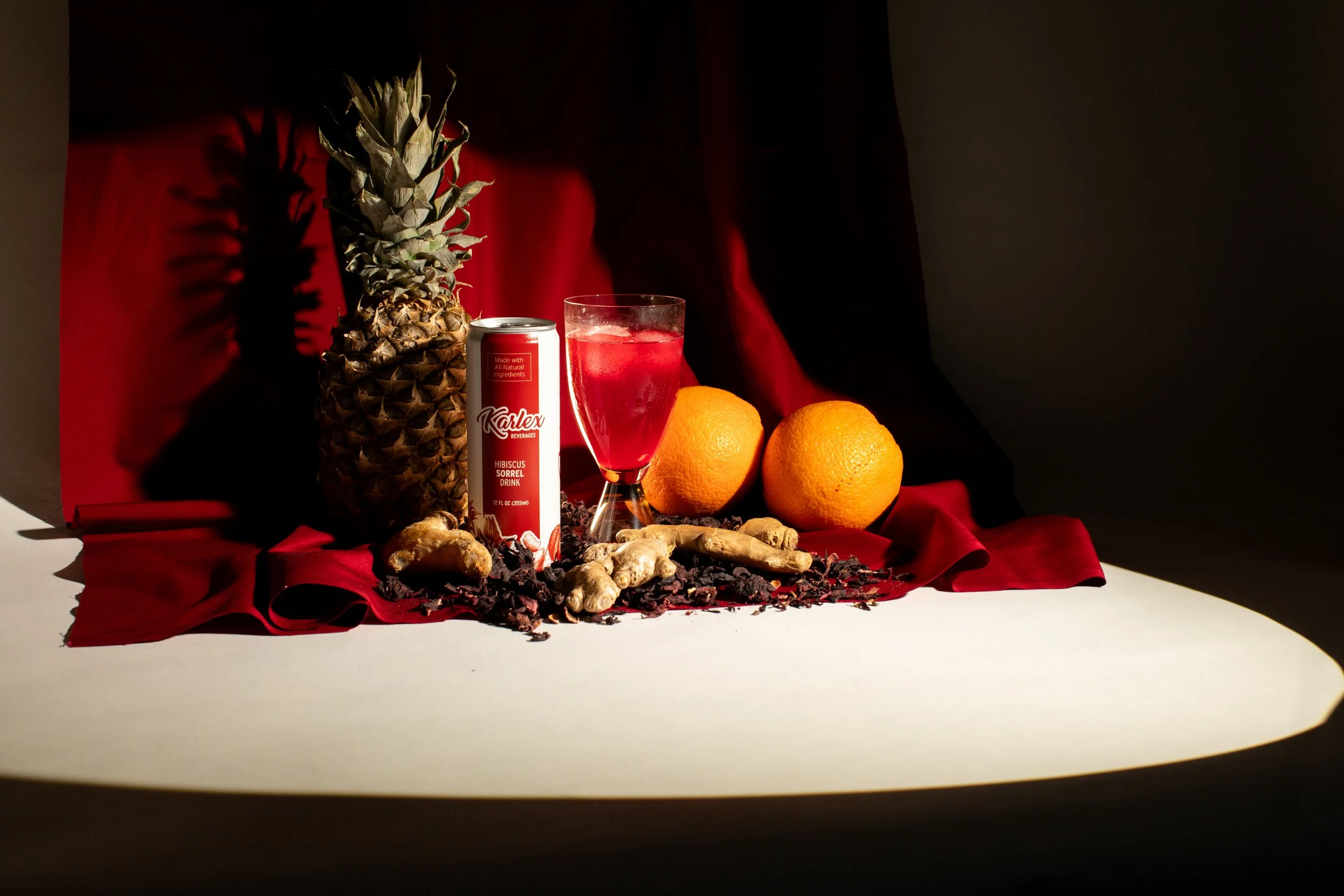 A still life arrangement featuring a pineapple, two oranges, a ginger root, a glass of red beverage, and a can of Karlex Hibiscus Sorrel Drink, all set against a red fabric backdrop with dramatic lighting.