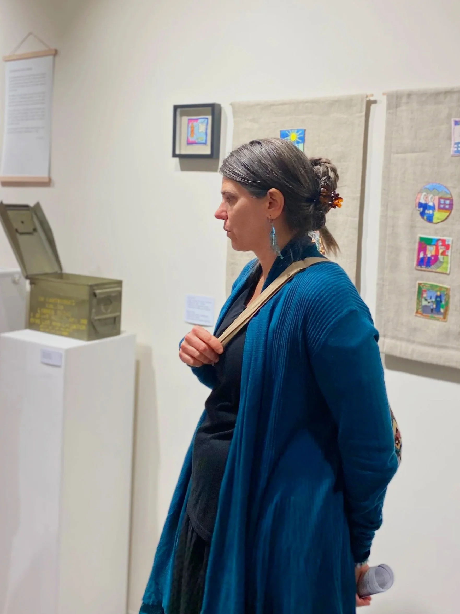 Seitenansicht einer Frau mit braunem Haar in einem Zopf, die in einem Kunstmuseum vor Wandbehängen mit kindlichen Illustrationen steht.