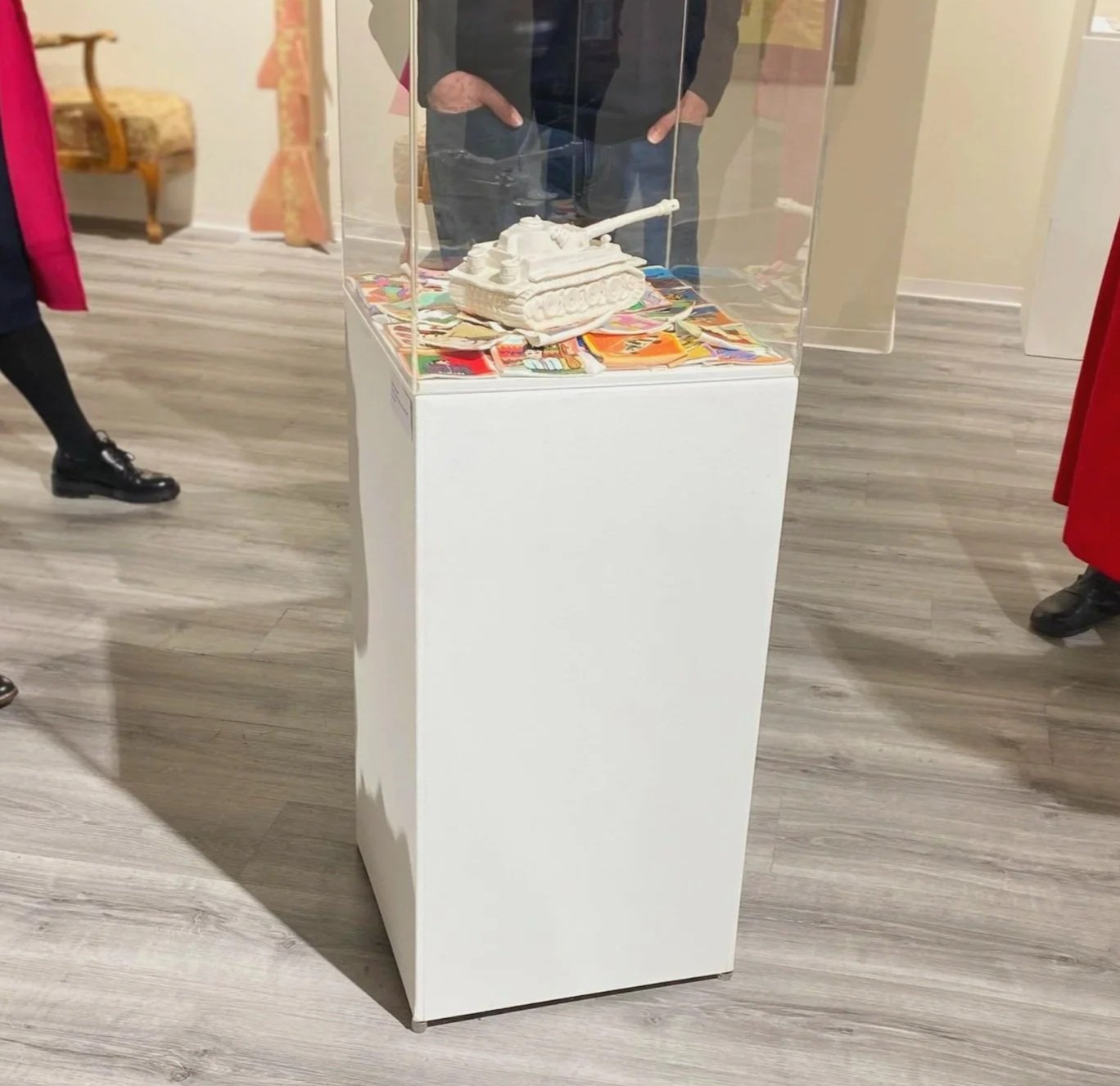 Kunstvoll gestaltete weiße Modellpanzer in einem transparenten Gehäuse auf einem weißen Podest, umgeben von bunten Kärtchen oder Postern in einem Museum oder einer Ausstellung.