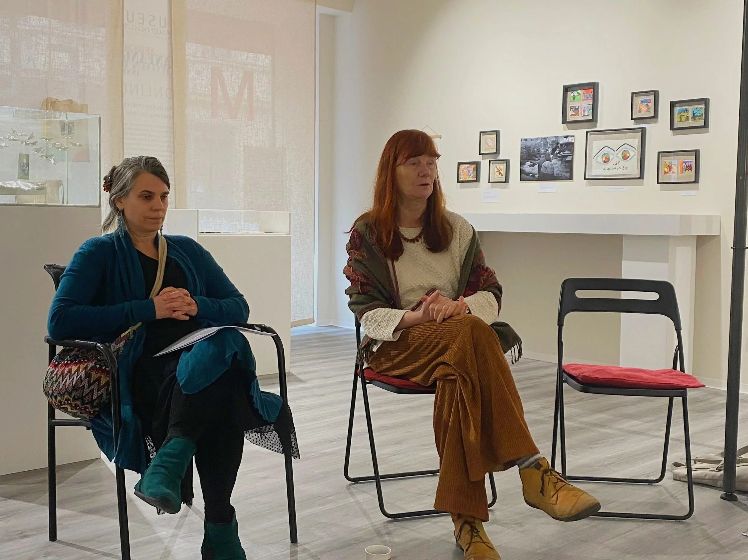 Zwei Frauen sitzen in einem Kunstgalerie, die eine Frau mit roten Haaren trägt braune Hosen, gelbe Stiefel, einen mehrfarbigen Pullover und eine Halskette. Die andere Frau mit grauem Haar trägt eine blaue Jacke, schwarze Hose und grüne Schuhe. An der Wand sind mehrere kleine Bilder in schwarzen Rahmen zu sehen.