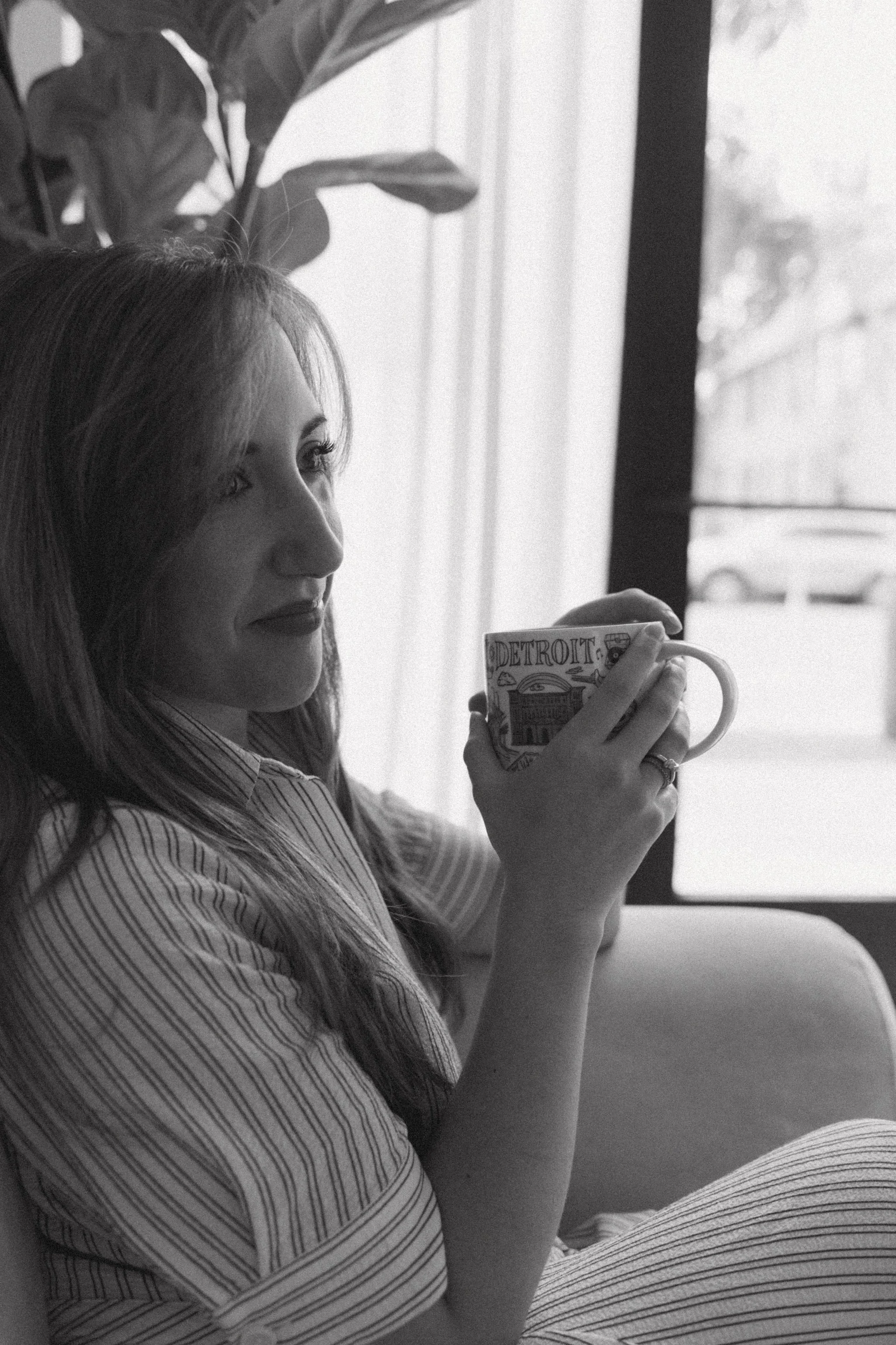 A woman with long hair and wearing a striped shirt sitting by a window, holding a mug with the word 'Detroit' on it, looking outside.