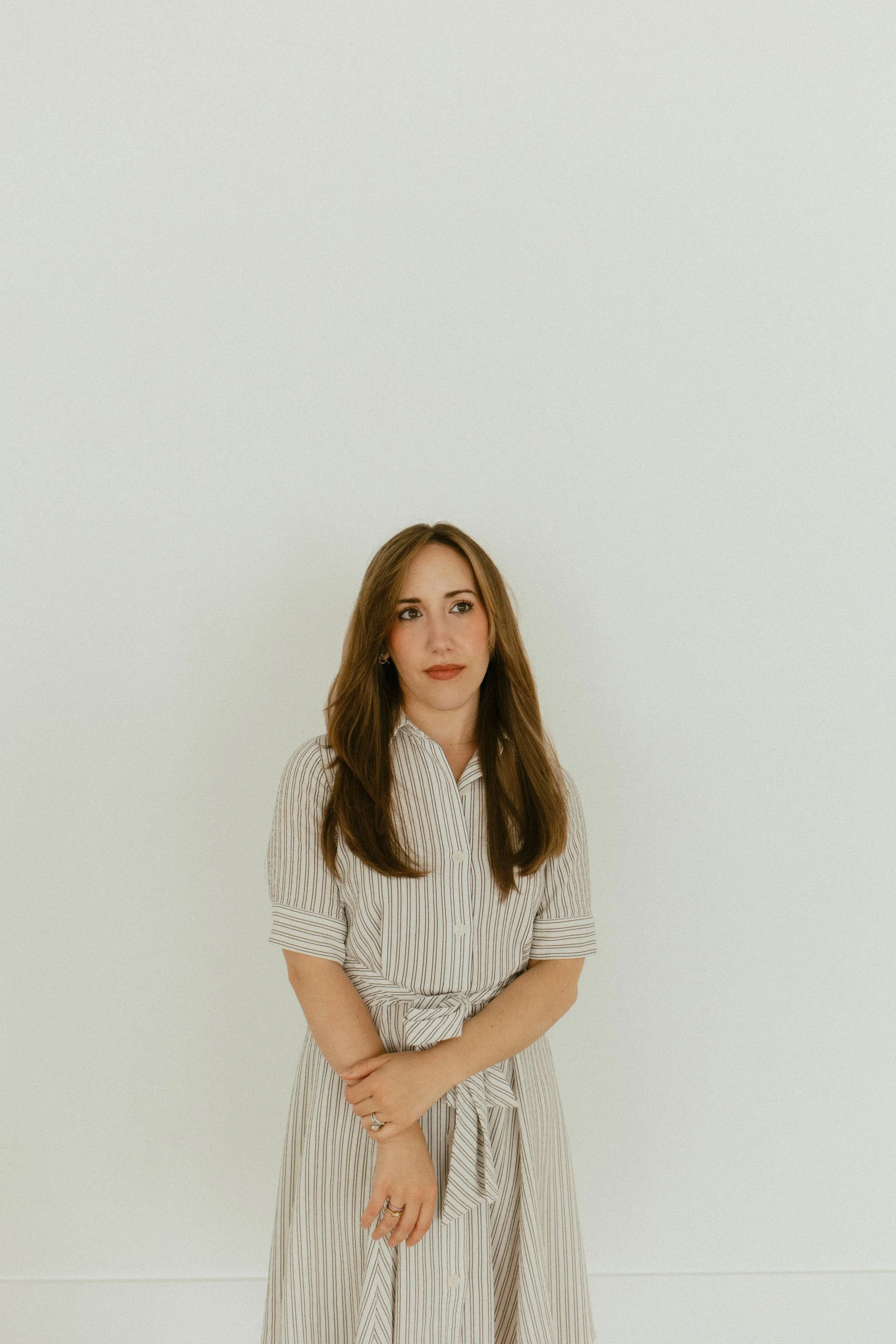 A woman with long brown hair wearing a striped beige and white dress standing against a plain white wall.