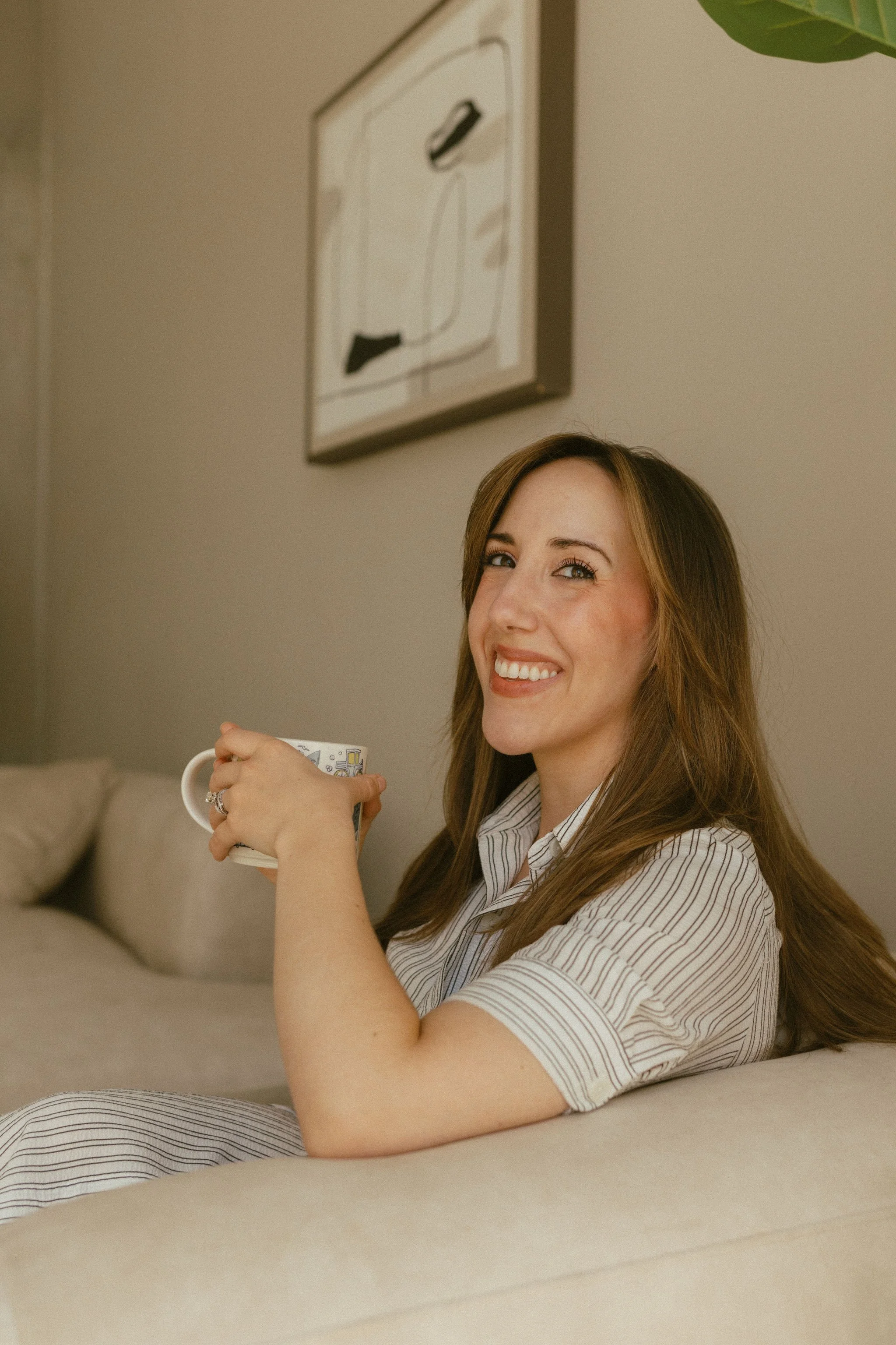 A woman with light brown hair smiling while holding a mug, sitting on a beige couch in a room with neutral-colored walls and artwork on the wall.