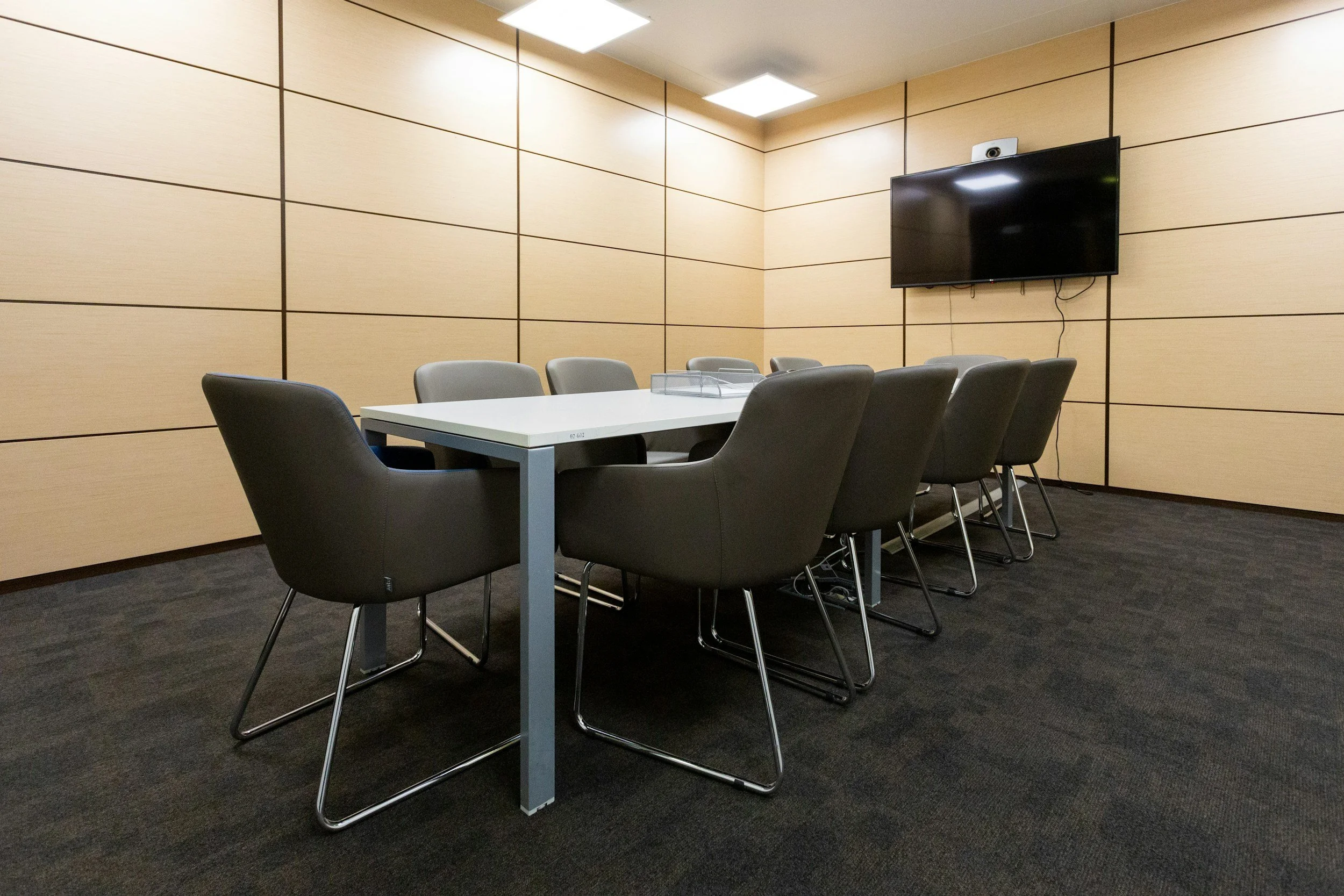 A modern conference room with a rectangular white table, eight black and gray chairs, beige wall panels, a wall-mounted flat-screen TV, and ceiling lights.