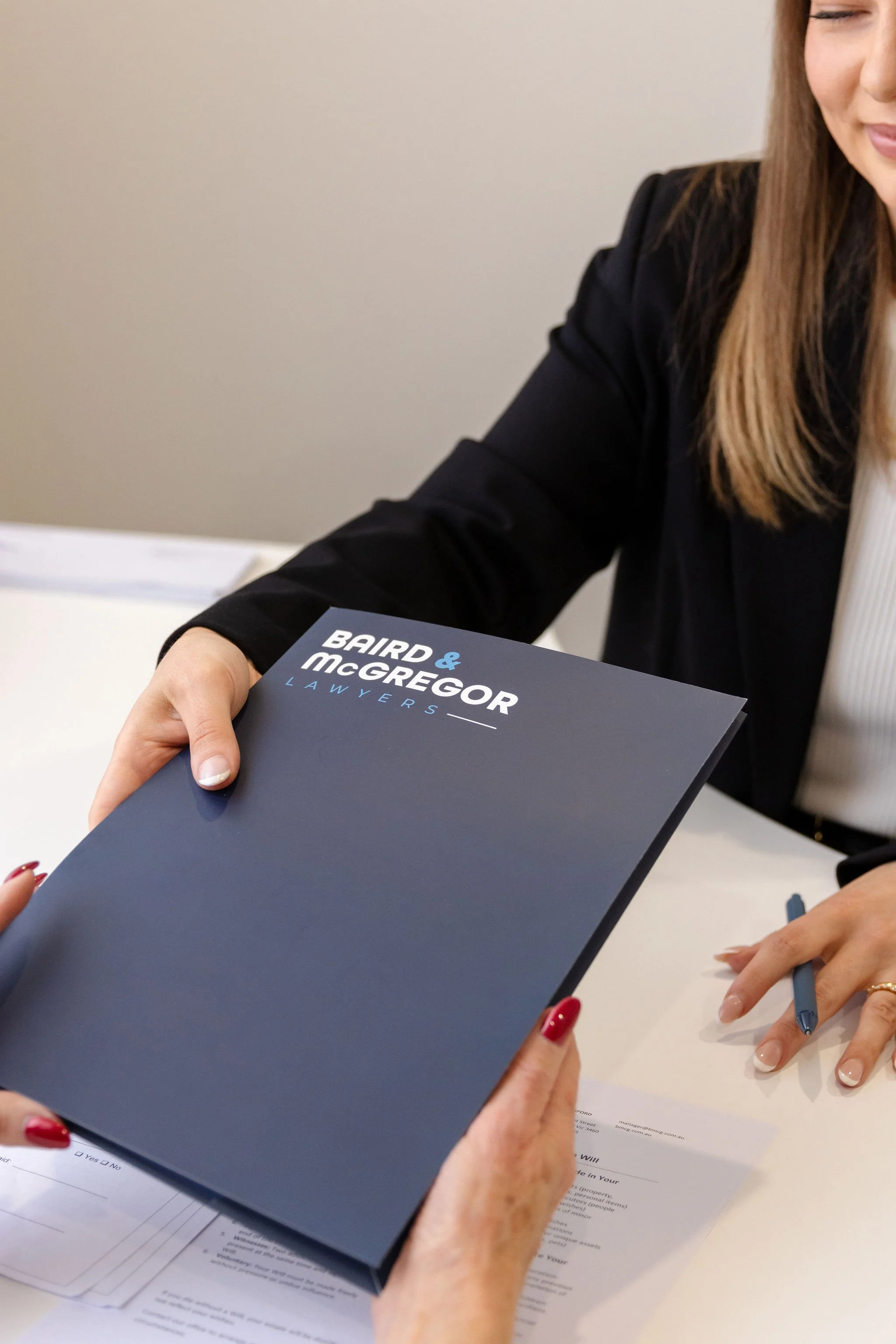 Close-up of two women exchanging a Baird & McGregor law firm brochure at a business meeting.