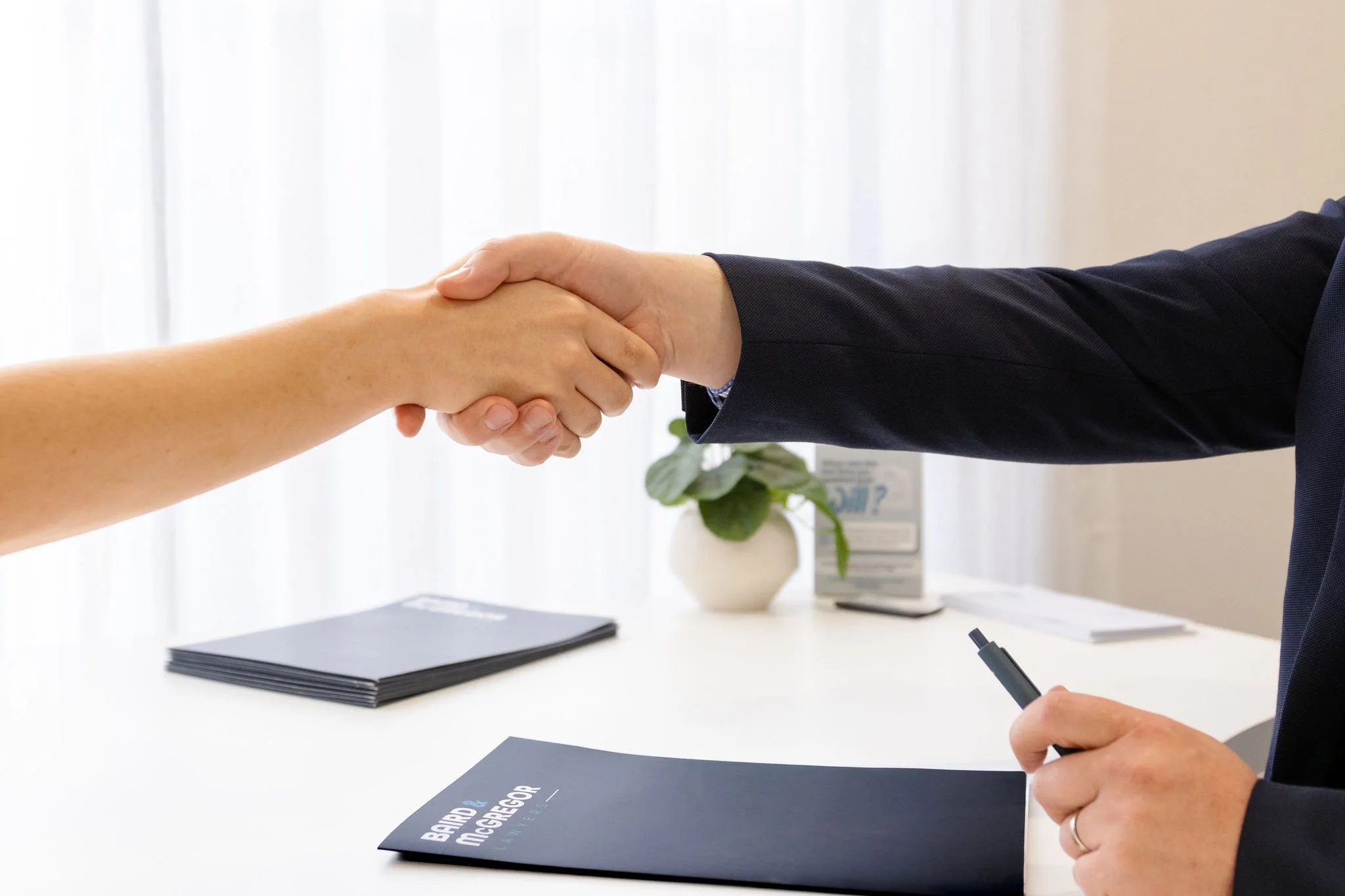 Two people shaking hands over a desk with notebooks and a pen, indicating a business agreement or deal.