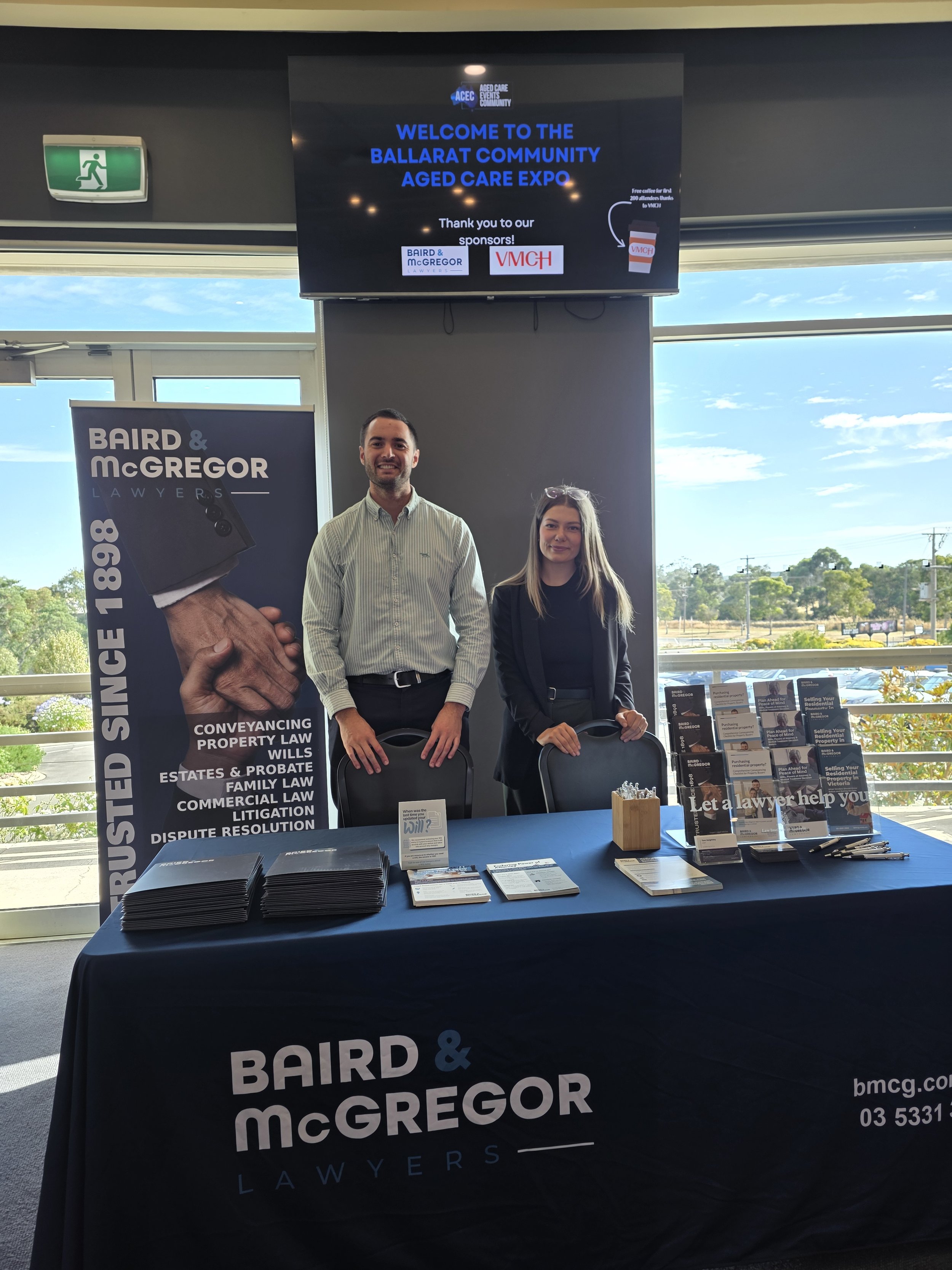 Two people standing behind a table at the Ballarat Community Aged Care Expo. The table displays brochures and pens, with a large Baird & McGregor Lawyers banner and a promotional sign. A screen behind shows the event's welcome message and sponsor logos.