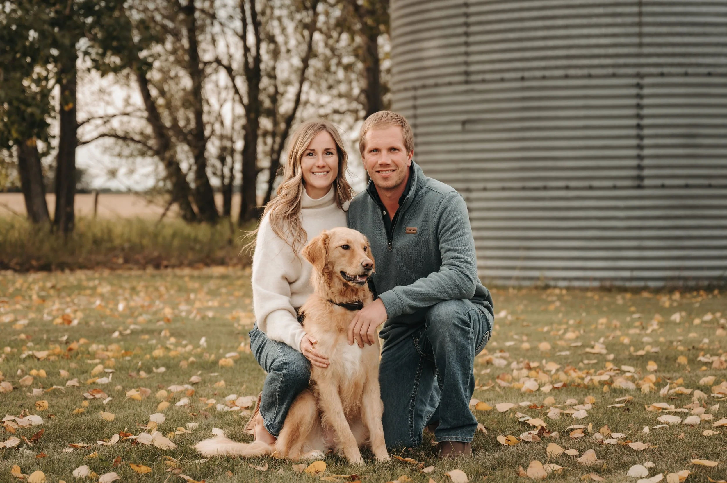 A smiling couple posing outdoors on a fall day with their golden retriever puppy, with trees and a silo in the background.