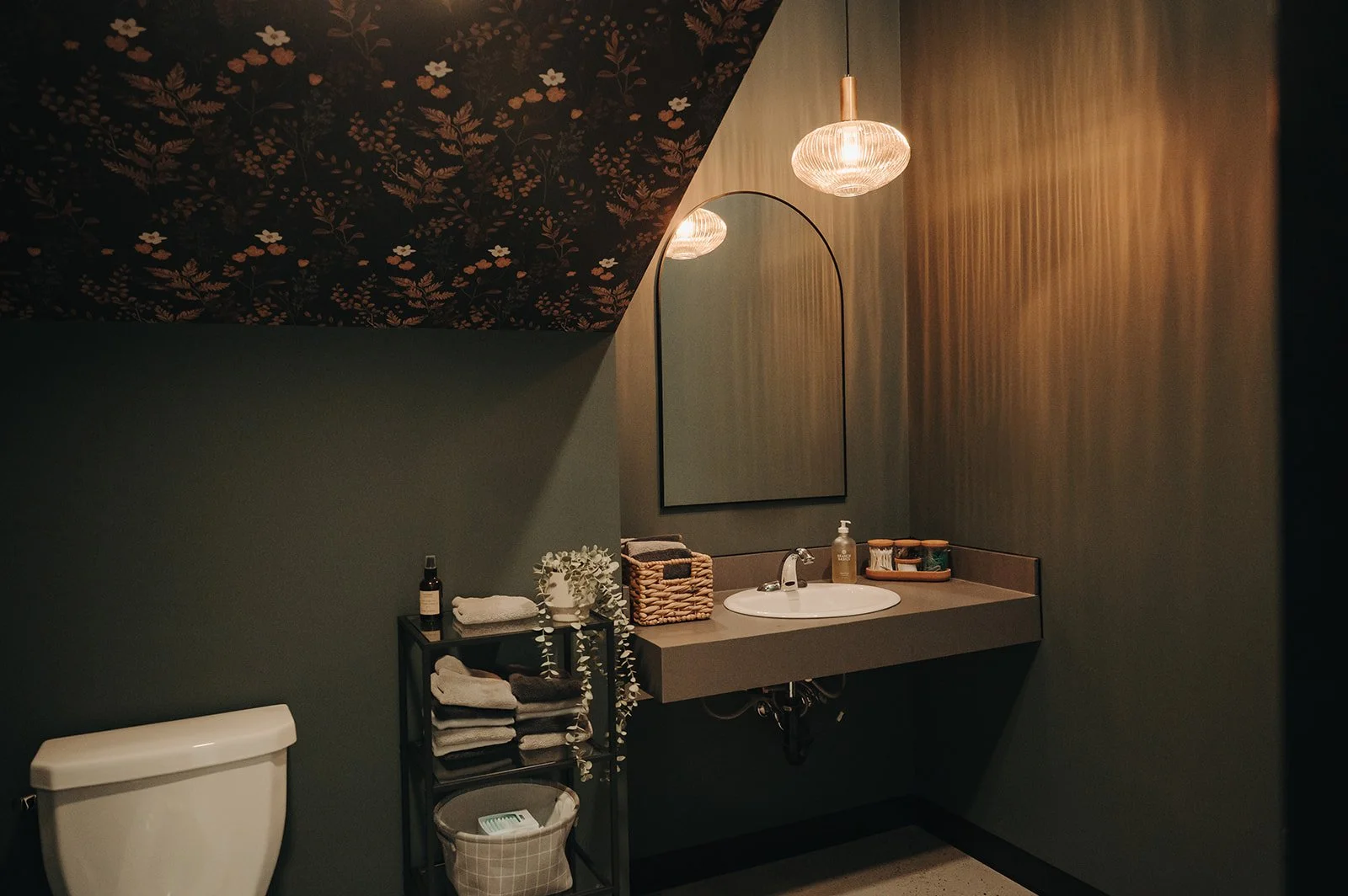 A bathroom with dark floral wallpaper on the ceiling, a large mirror, a floating sink with a bottle of hand soap, a wicker basket, folded towels, a small black shelving unit with toiletries and towels, and soft lighting from two pendant lights.