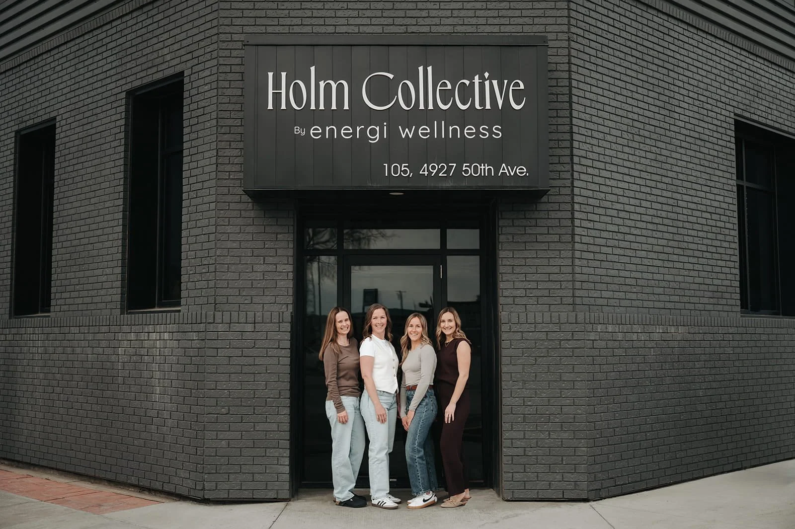 Four women standing outside a black brick building with a sign that reads "Holm Collective by energi wellness, 105, 4927 50th Ave."