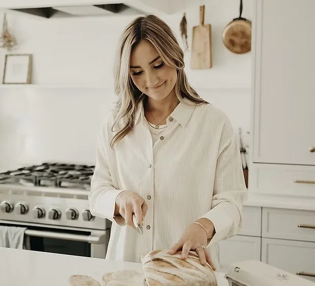 A woman with long, wavy hair cutting a loaf of bread in a modern kitchen with white cabinets and a stove.
