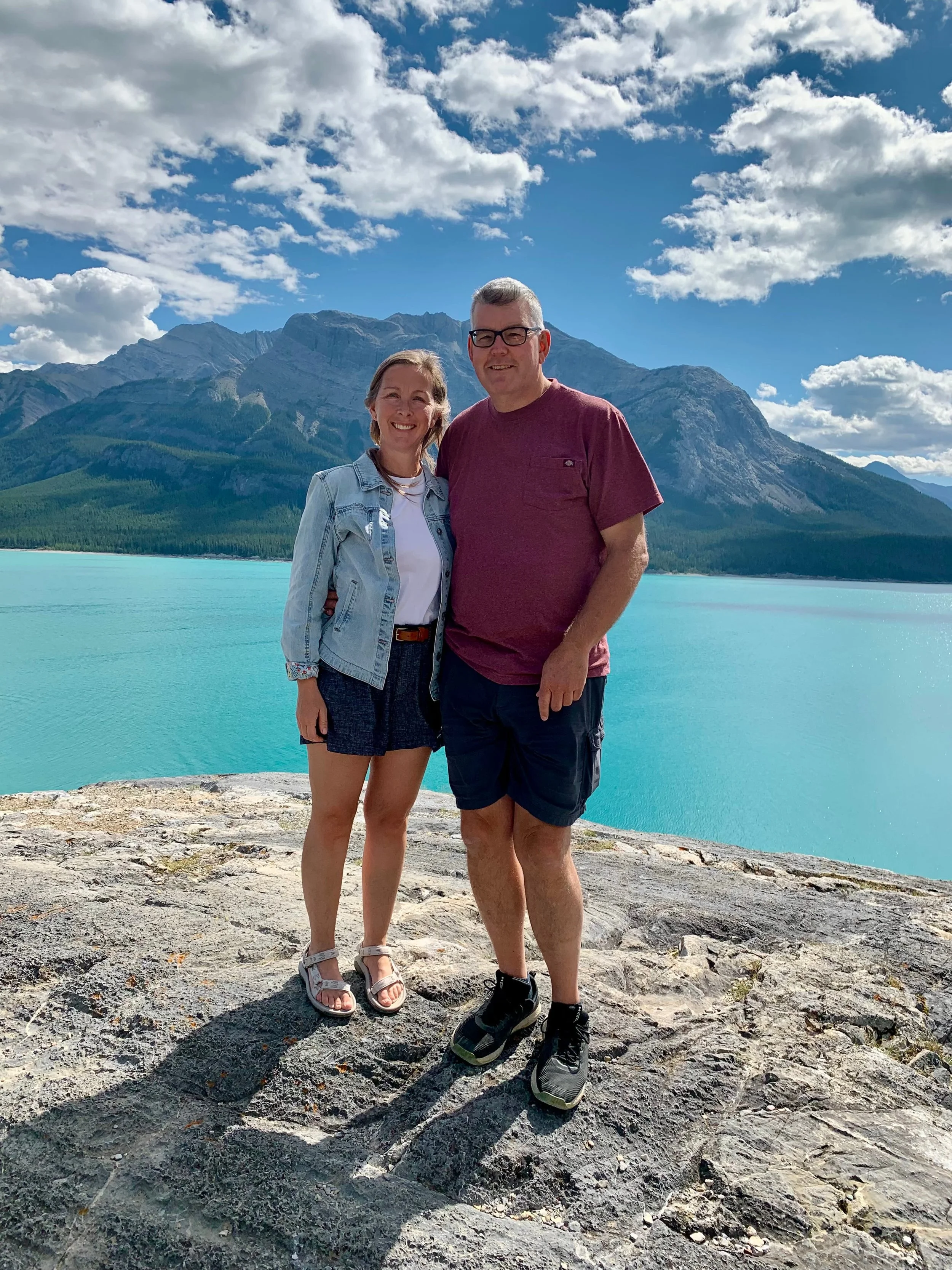 A smiling woman and man standing on a rocky area near a body of turquoise water with mountains and partly cloudy sky in the background.