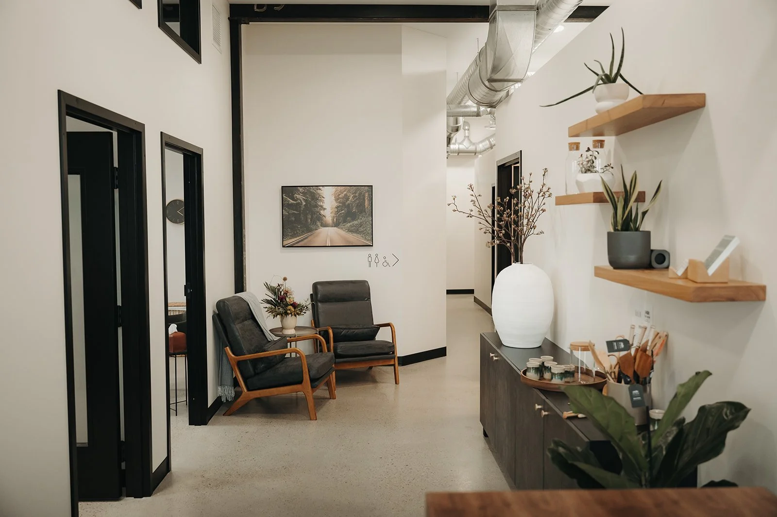 A modern waiting area with two black chairs featuring wooden frames, a small table with flowers, a large white vase with branches, wall-mounted shelves with decorative items, at Holm Collective, Lacombe AB.