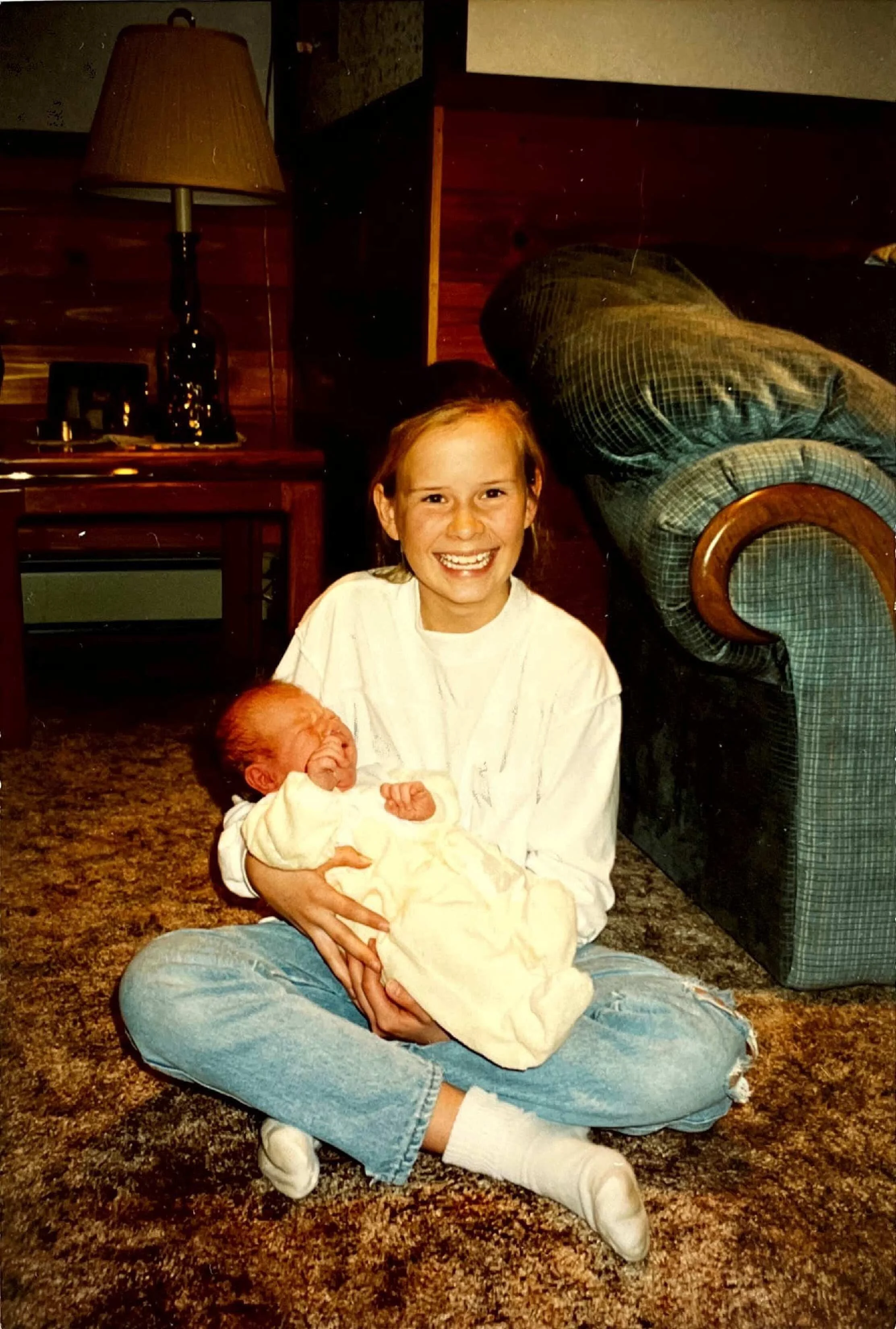 A young girl with red hair and a big smile sitting on a brown patterned carpet, wearing a white shirt, blue jeans, and white socks, holding a newborn baby wrapped in a cream-colored blanket. Behind them, there is a wooden side table with a lamp, a picture frame, and other items, and an armchair with plaid upholstery.