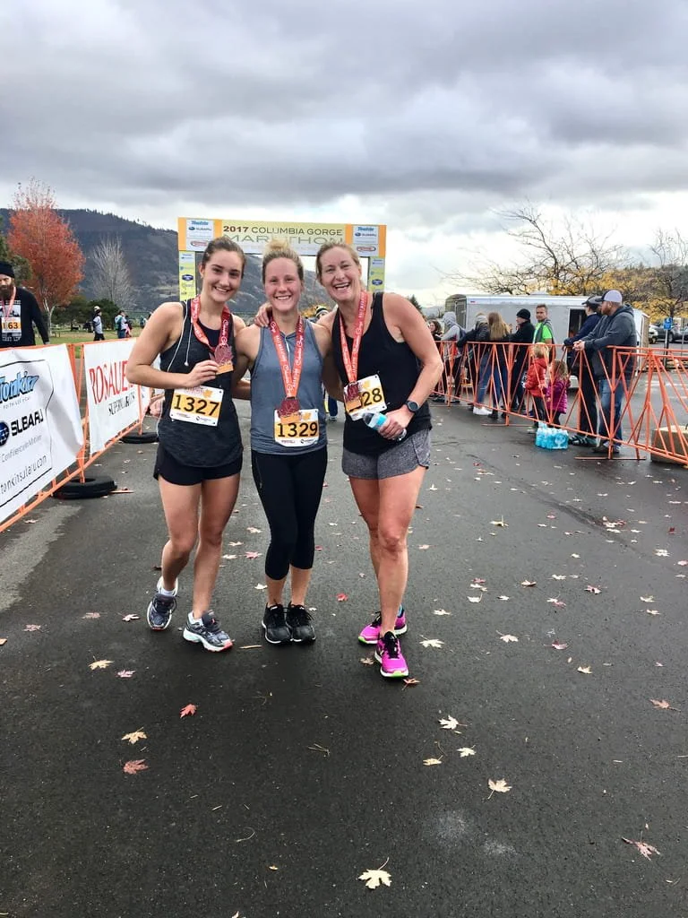 Three women wearing race bibs and medals, smiling and posing together after finishing a race at the 2017 Columbia Gorge Half Marathon, on a grey cloudy day, with spectators and race barricades in the background.